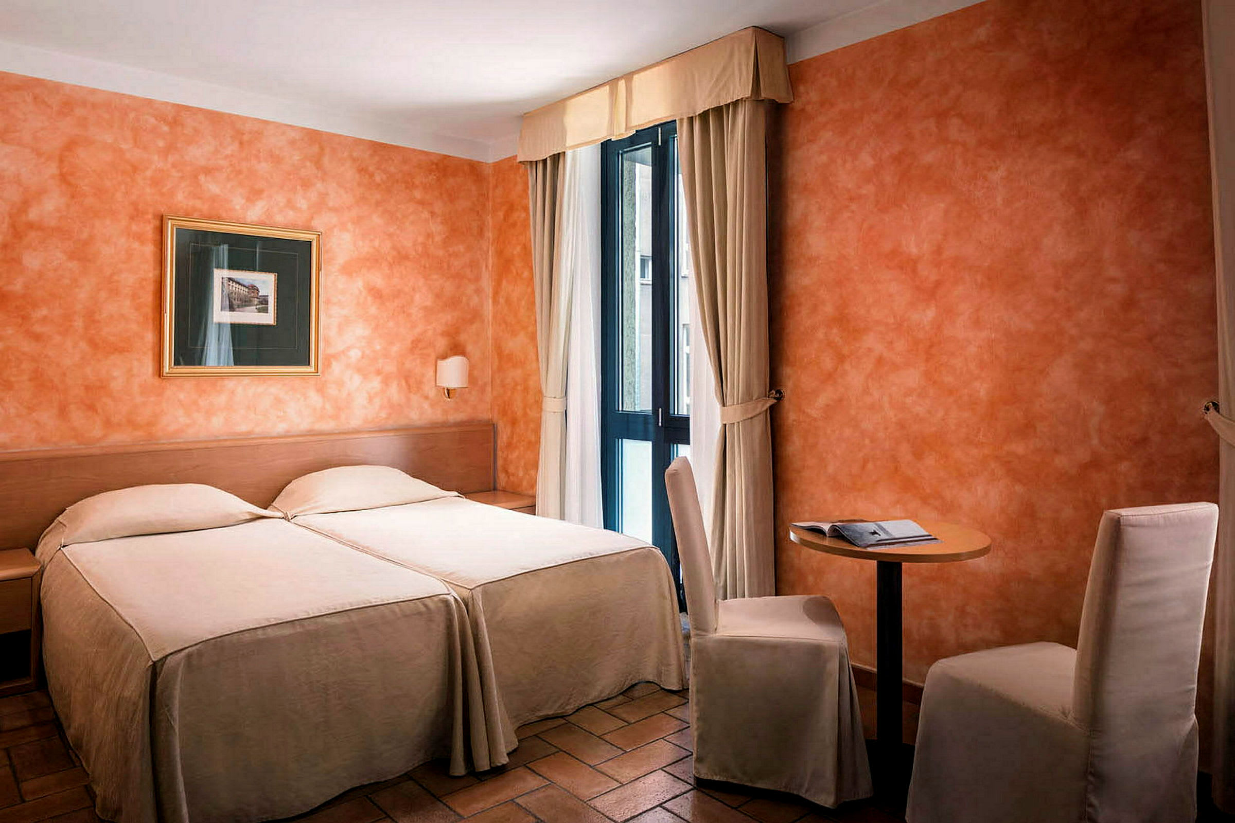 Et hotellrom på bildet med senger i lyse trekk, oransje vegger, et lite bord med to stoler og et maleri. Vinduer med lyse gardiner slipper inn dagslys. (AI-generert tekst)