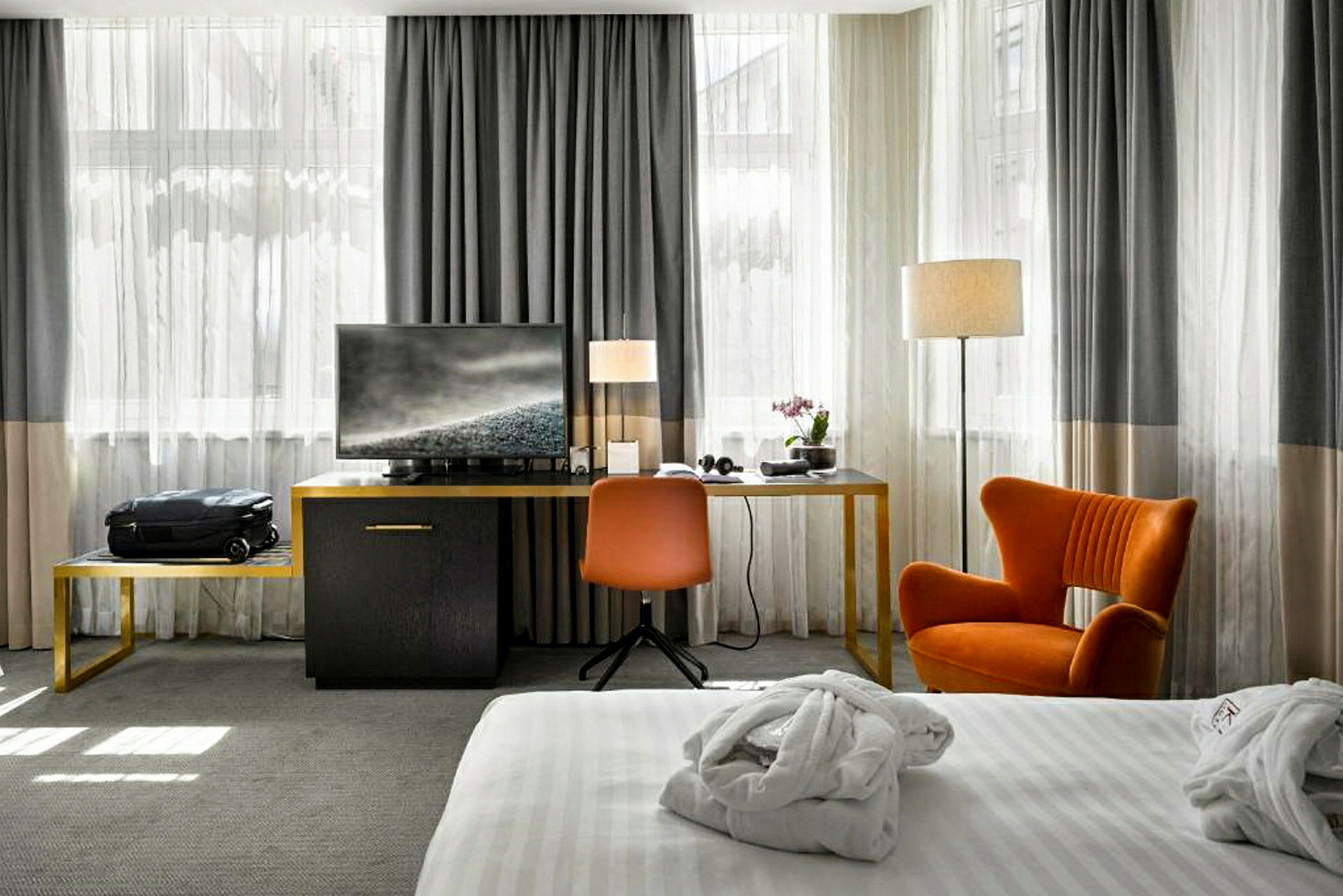 Et hotellrom med en seng med hvitt sengetøy i forgrunnen, og en sittegruppe med oransje stol og skrivebord med en TV i bakgrunnen. (AI-generert tekst)