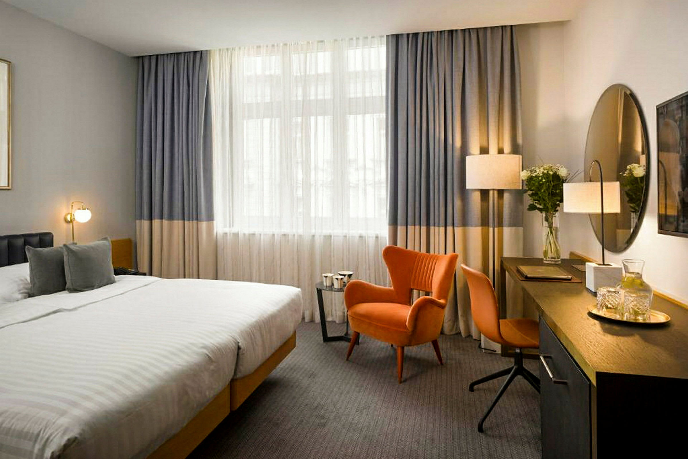 Et hotellrom med en stor seng, en oransje lenestol og skrivebord med speil. (AI-generert tekst)