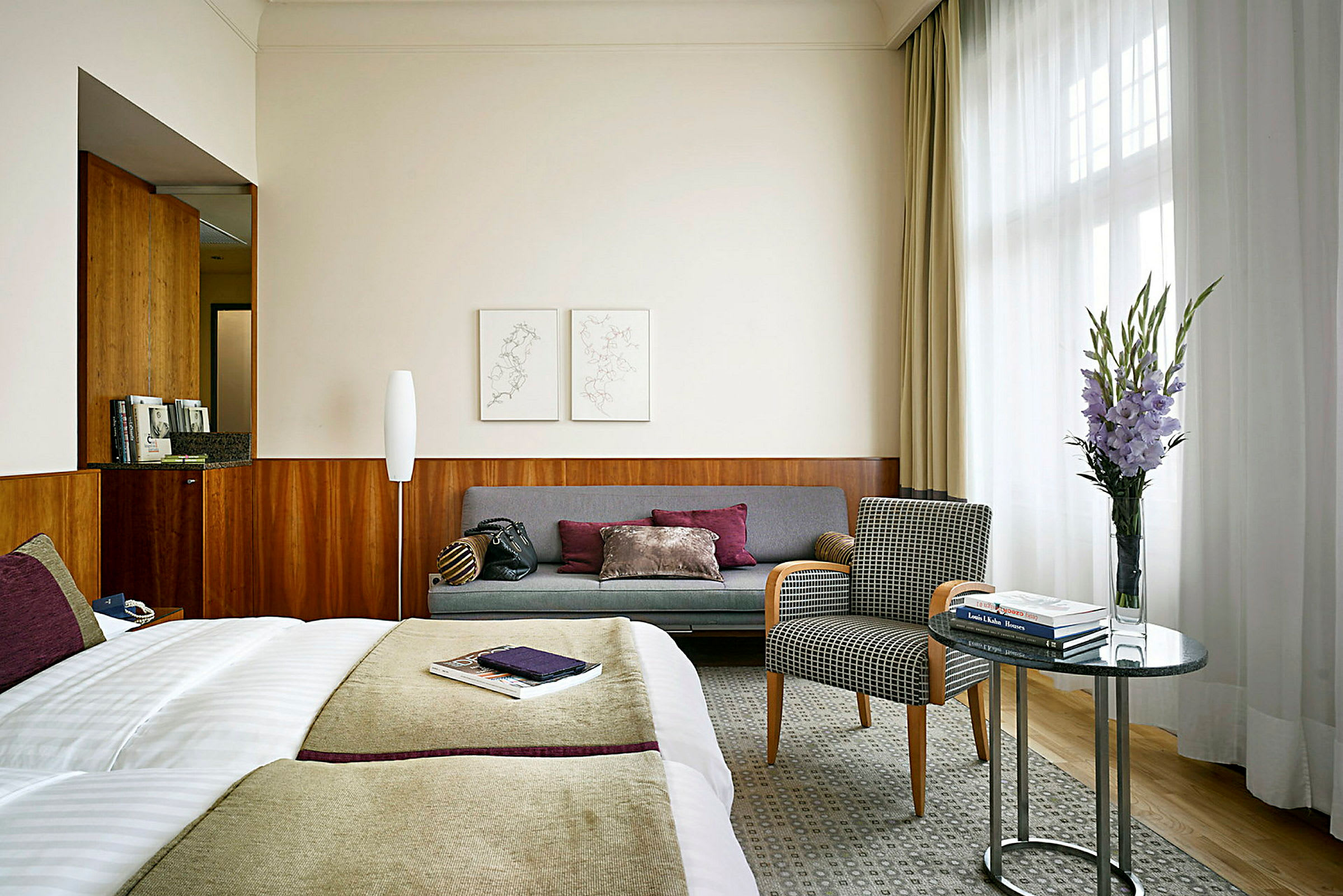 Et hotellrom med en seng, en sofa, en lenestol og et bord med en vase med blomster. (AI-generert tekst)