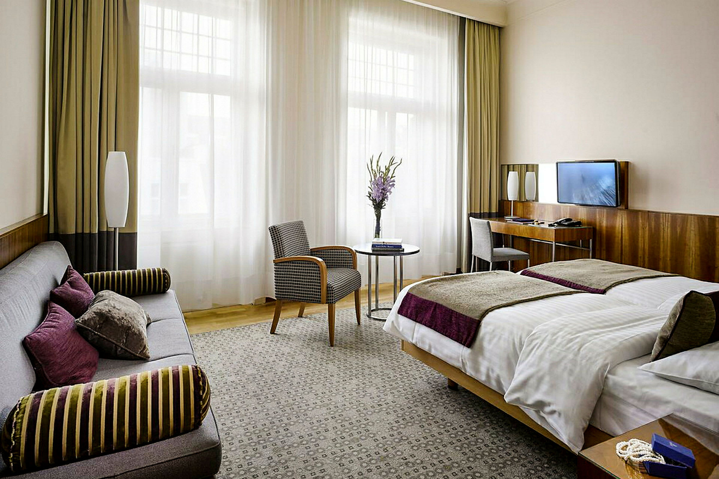 Et hotellrom med en dobbeltseng, sofa, lenestol og bord. Store vinduer med gardiner slipper inn naturlig lys. (AI-generert tekst)