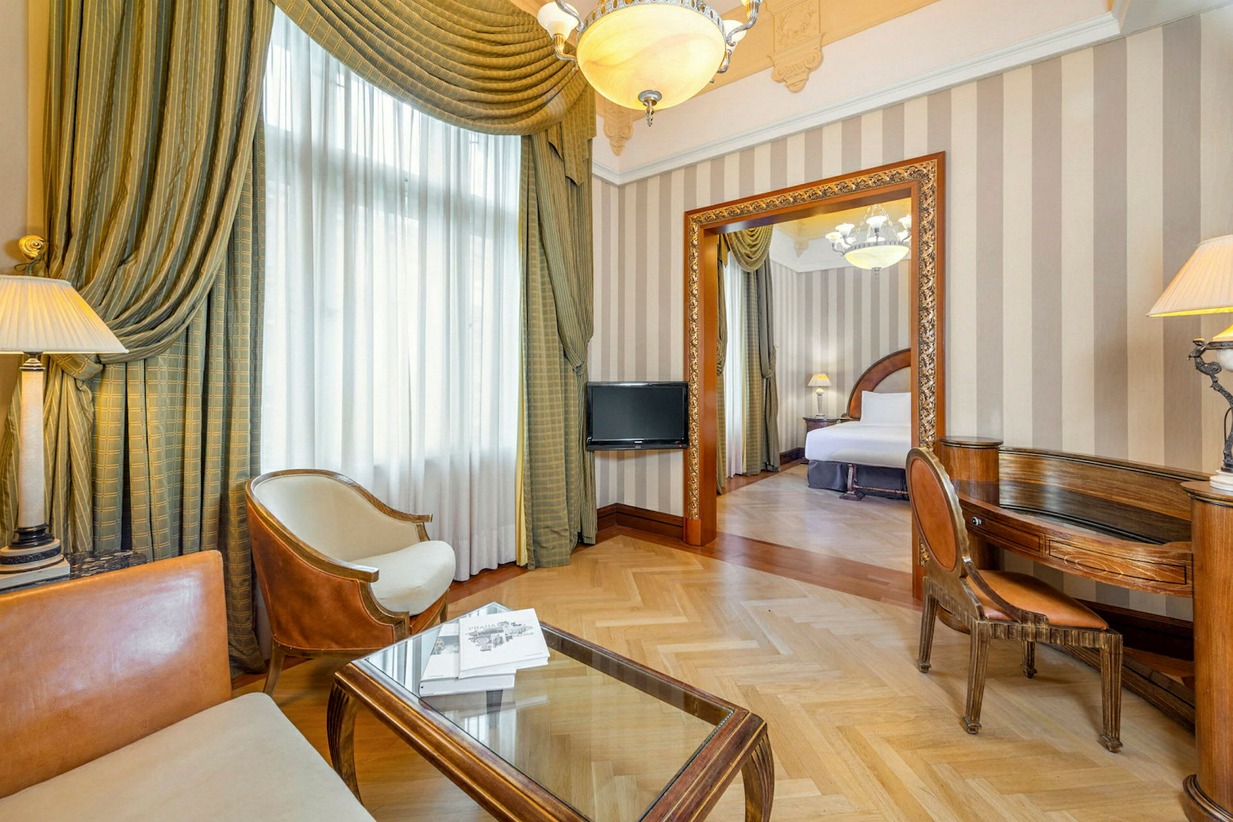 Et elegant hotellrom med en sittegruppe bestående av stoler og bord i forgrunnen, samt en åpen dør til soverommet med en bred seng. (AI-generert tekst)