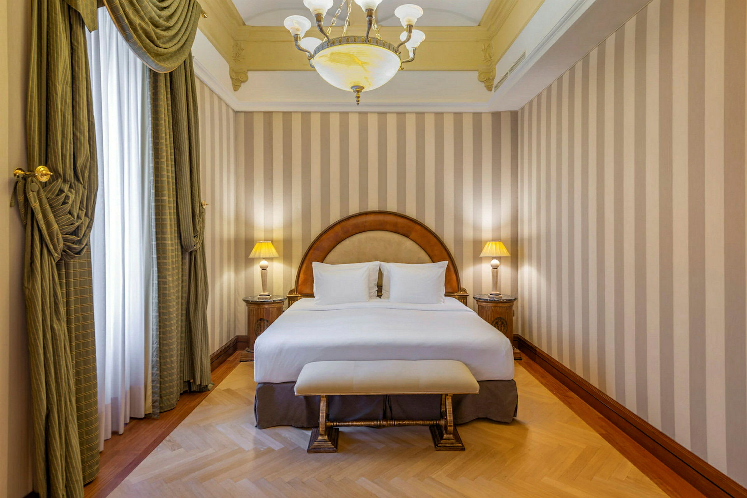Et hotellrom med en dobbeltseng, to nattbord med lamper, og elegante gardiner foran vinduet. (AI-generert tekst)
