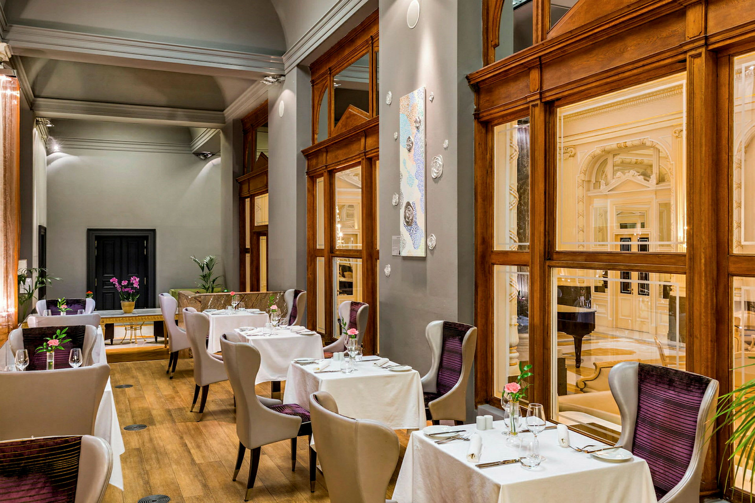 Bildet viser en elegant innredet restaurant med moderne stoler og bord kledd med hvite duker. Store vinduer og tredetaljer gir et luksuriøst inntrykk. (AI-generert tekst)