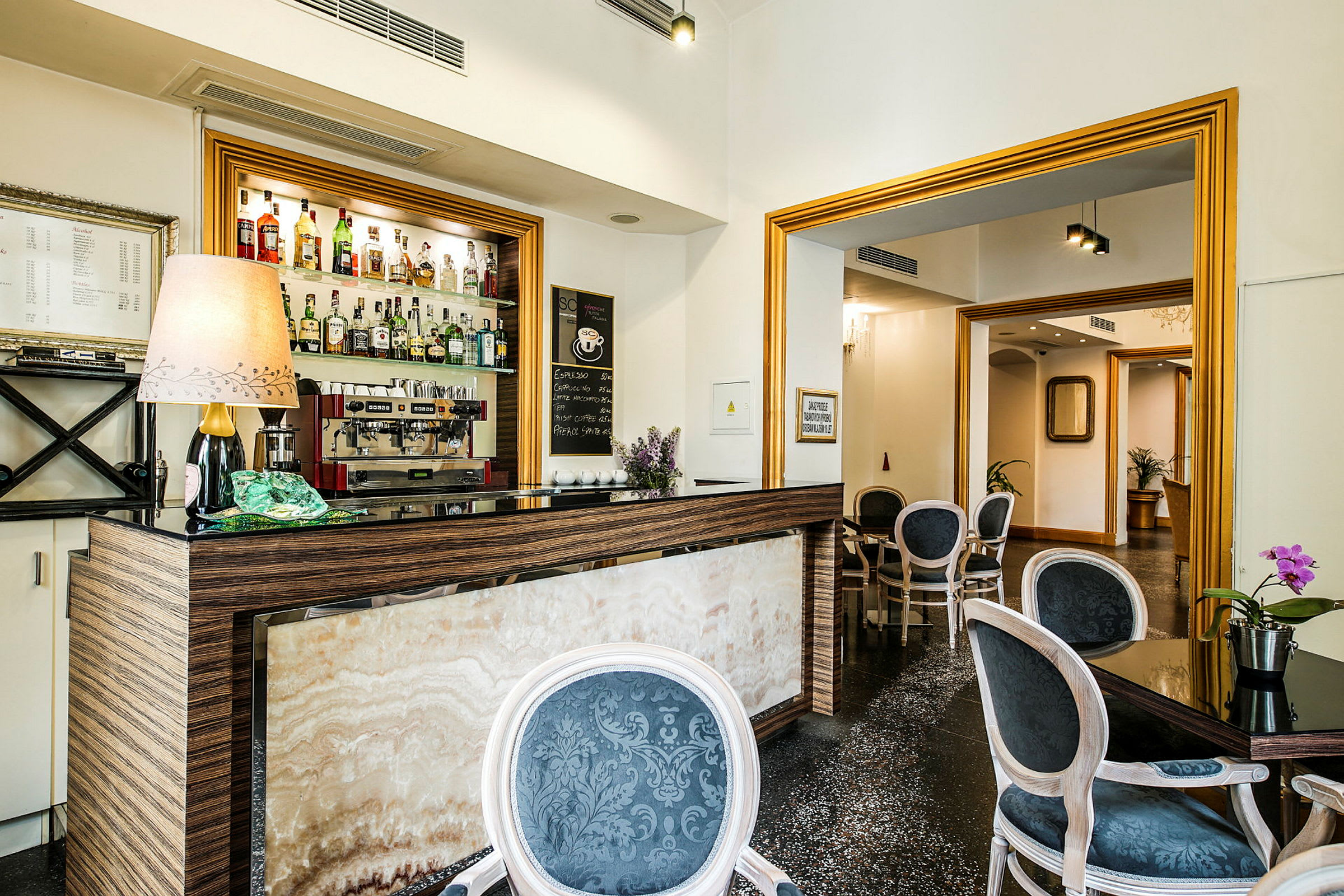 Bilde av en liten hotellbar med forskjellige typer brennevin på hyllen samt en kaffemaskin, omgitt av møblerte sitteplasser. (AI-generert tekst)