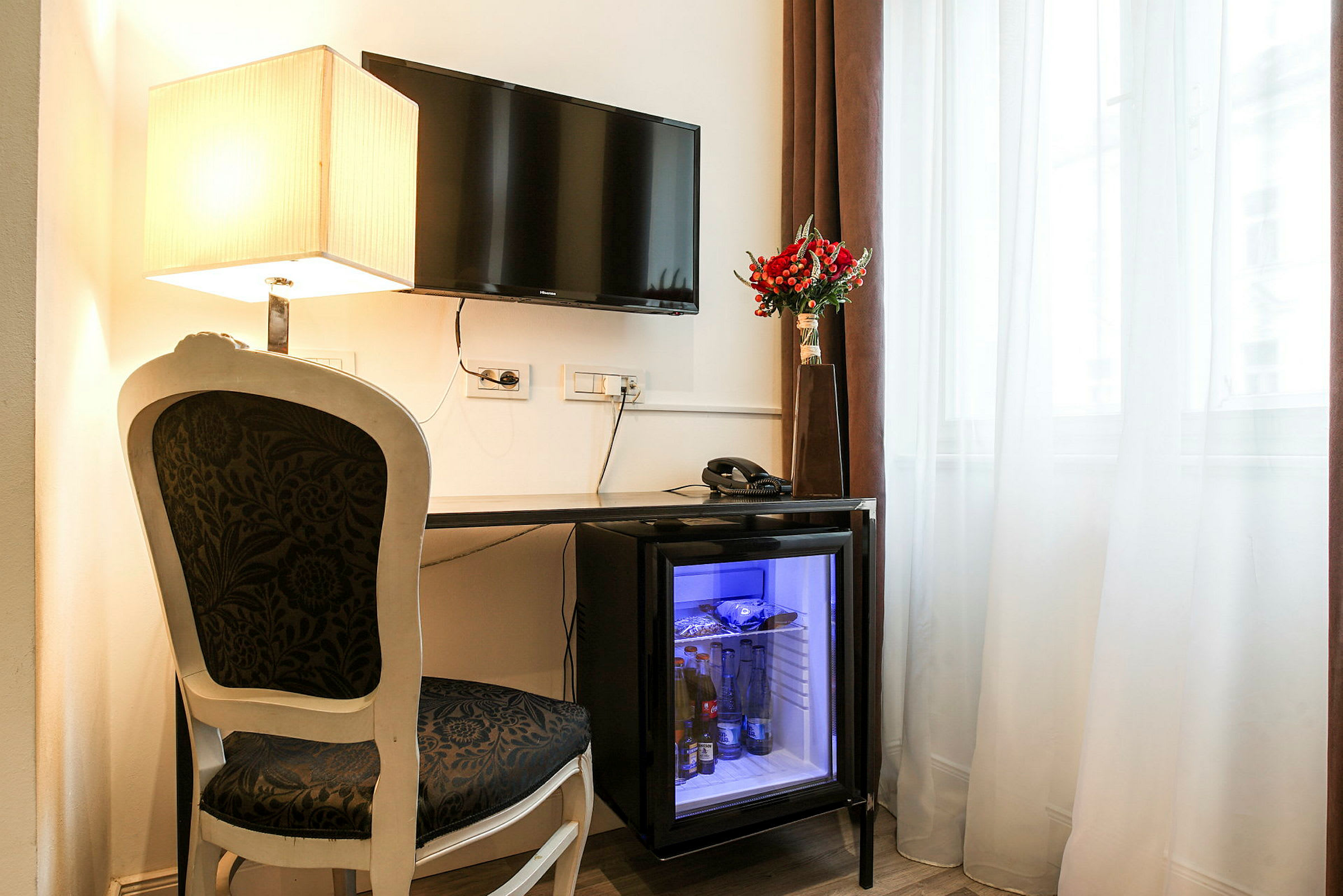 En skrivebordsområde med en stol og et bord. Bordet har en minibar med glassdør, en telefon og en vase med røde blomster. Et vindu med gardiner og en veggmontert TV samt en bordlampe. (AI-generert tekst)