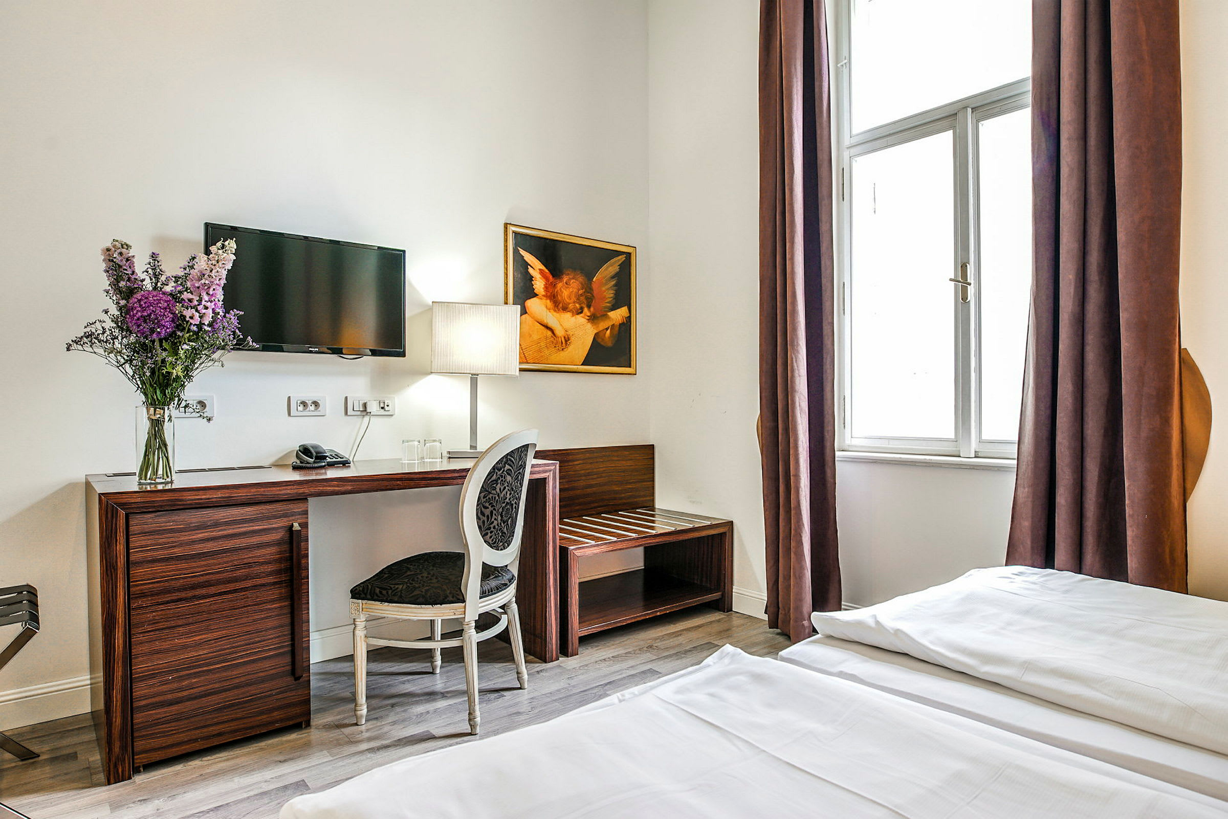 Et hotellrom med et skrivebord og stol, en TV på veggen, en blomsterbukett og en lampe plassert på bordet, samt et stort vindu med brune gardiner. (AI-generert tekst)