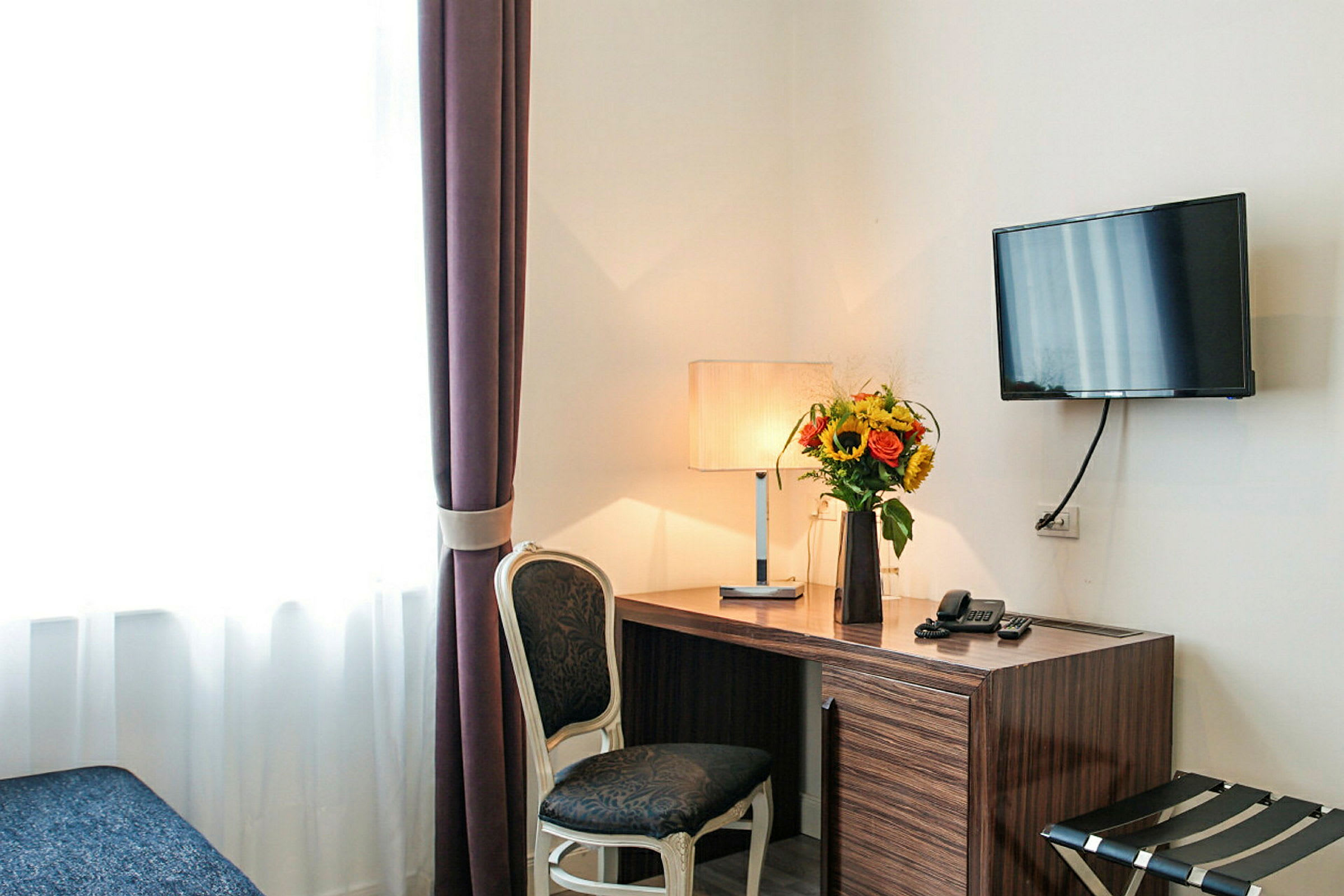 Et skrivebord med en lampe og en blomsterbukett på toppen, ved siden av en stol og en veggmontert TV. (AI-generert tekst)