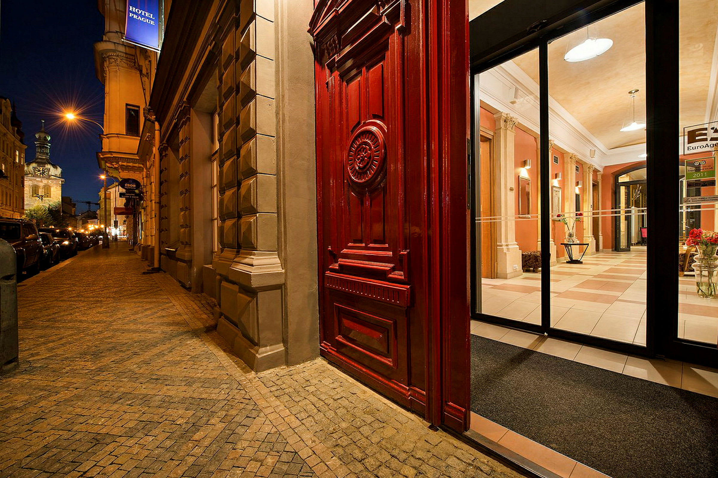 Inngang med en stor rød dør og glassdører som fører inn til et opplyst hotell med elegant interiør. (AI-generert tekst)