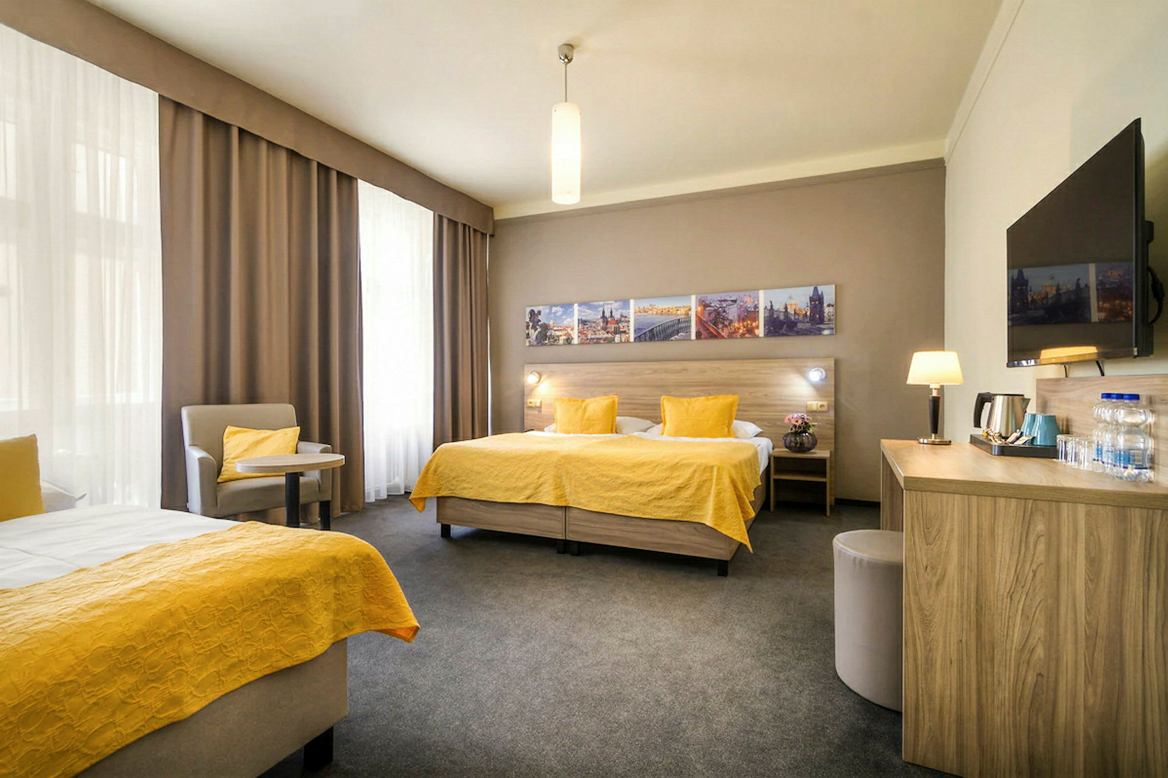 Et hotellrom med to enkeltsenger med gule sengetepper, en sittegruppe, et skrivebord og en veggmontert TV. (AI-generert tekst)