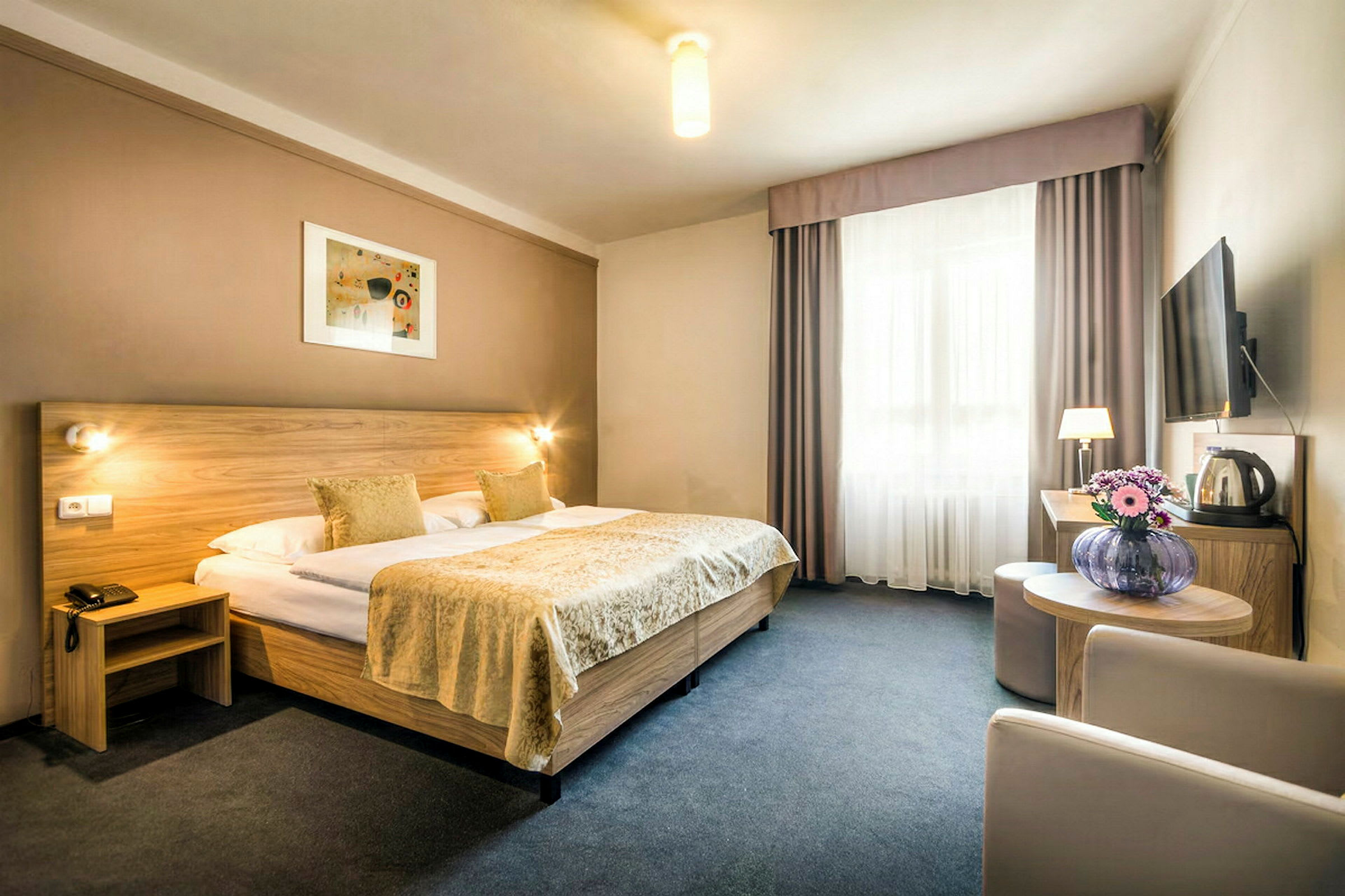 Et hotellrom med en dobbeltseng, en sittegruppe, blomster på et bord og en veggmontert TV. (AI-generert tekst)