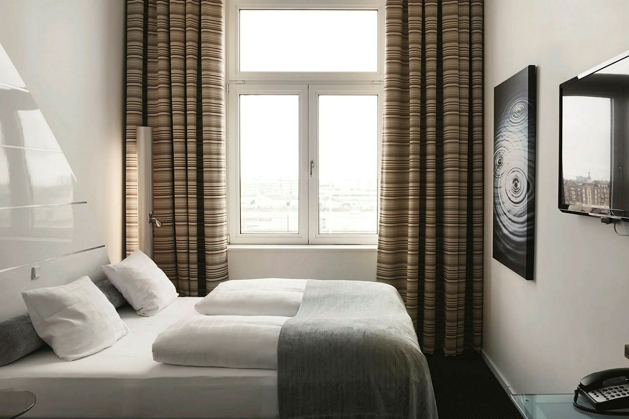 Hotellrom med seng, store vinduer, stripete gardiner og vegg-TV. (AI-generert tekst)