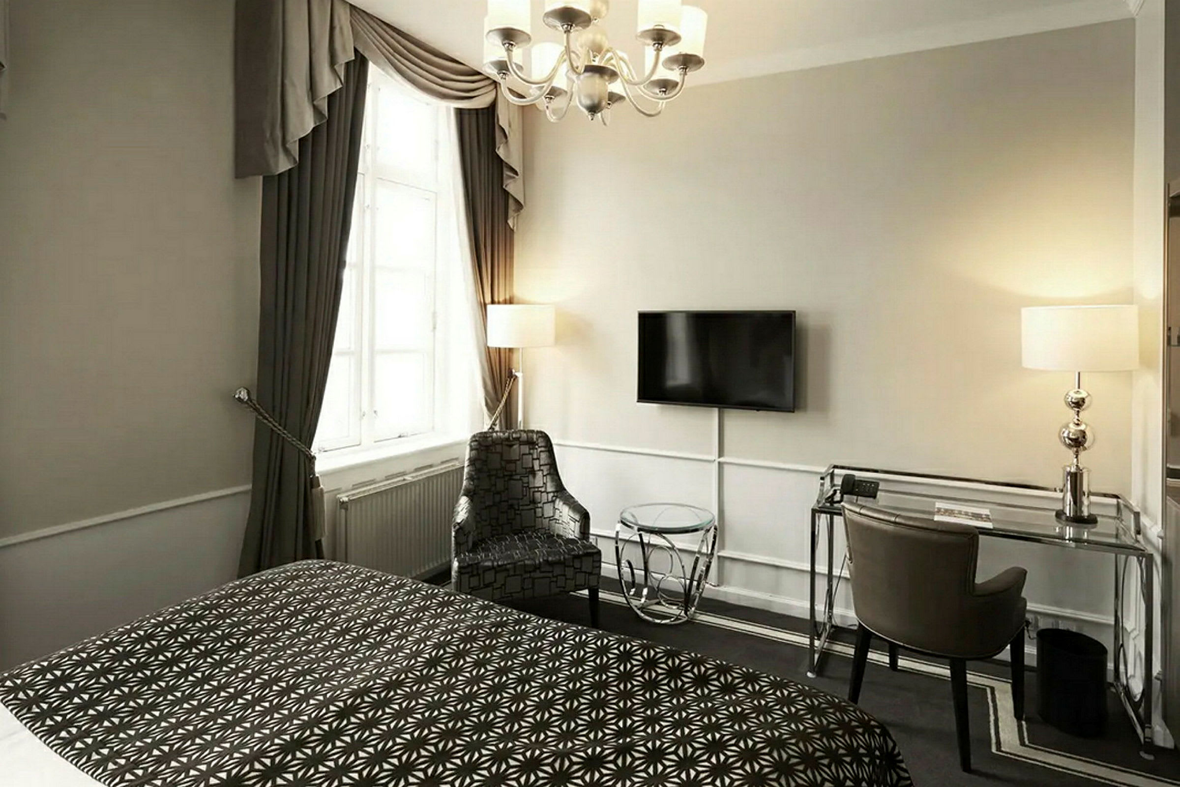 Et hotellrom med en seng, et skrivebord, en stol og en veggmontert TV. Et vindu med gardiner slipper inn dagslys. (AI-generert tekst)