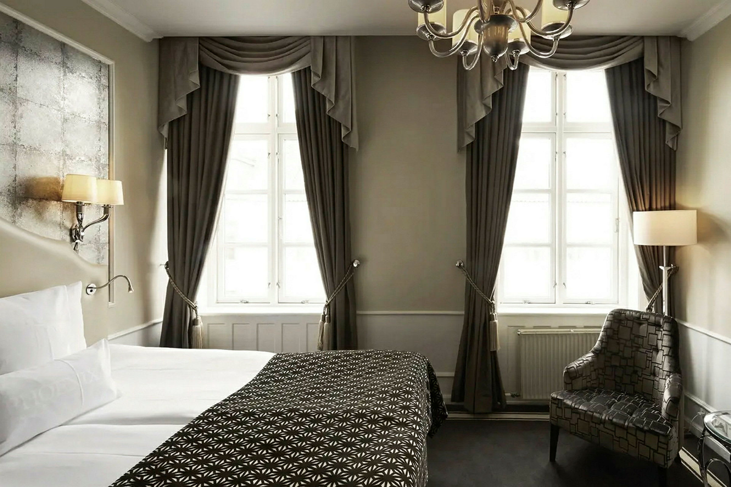 Et elegant hotellrom med en oppredd seng, store vinduer med mørke gardiner, en lenestol og en dekorativ taklampe. (AI-generert tekst)