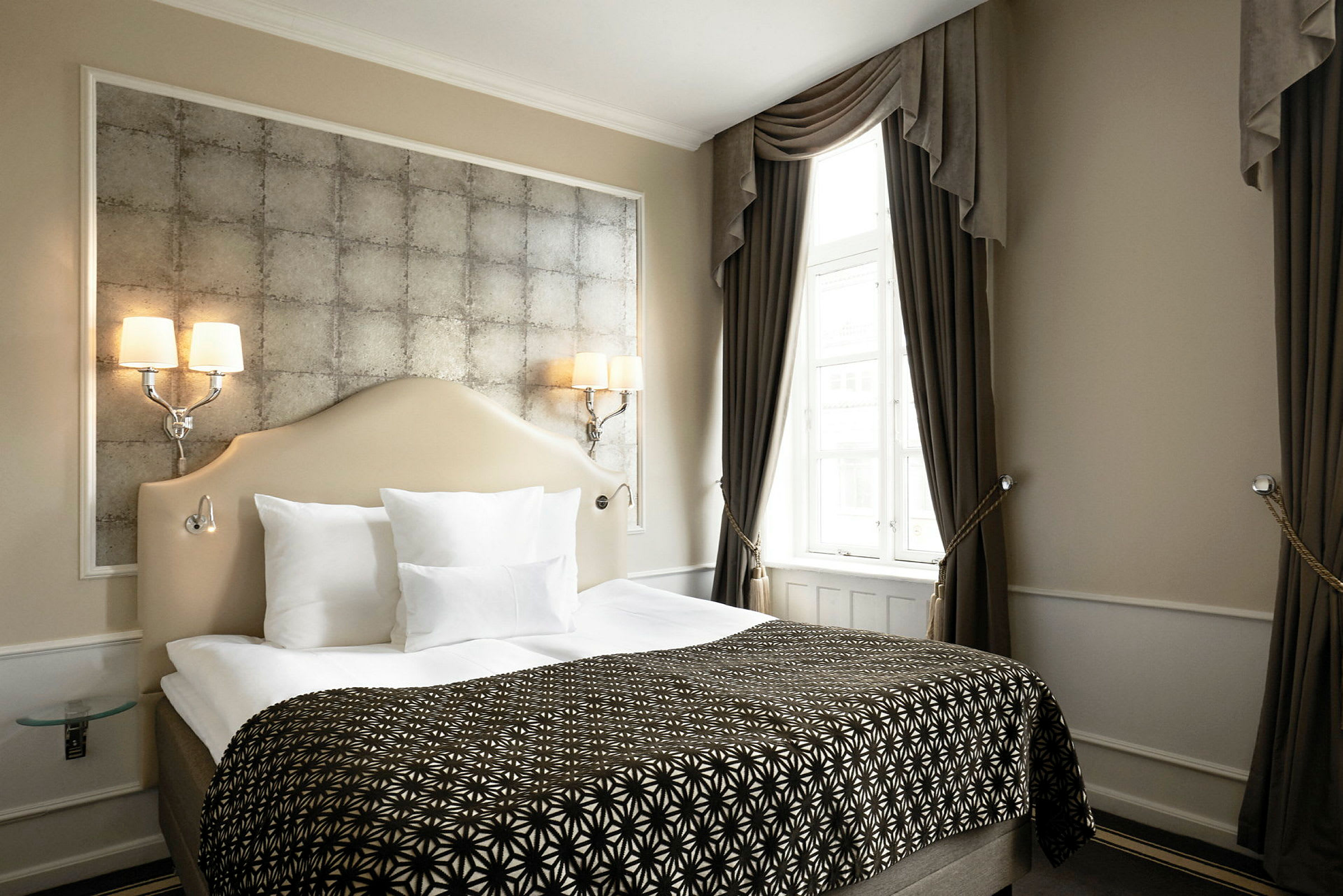 Et elegant hotellrom med en dobbeltseng, dekorative puter og smakfull innredning. Gardiner rammer inn et lyst vindu. (AI-generert tekst)