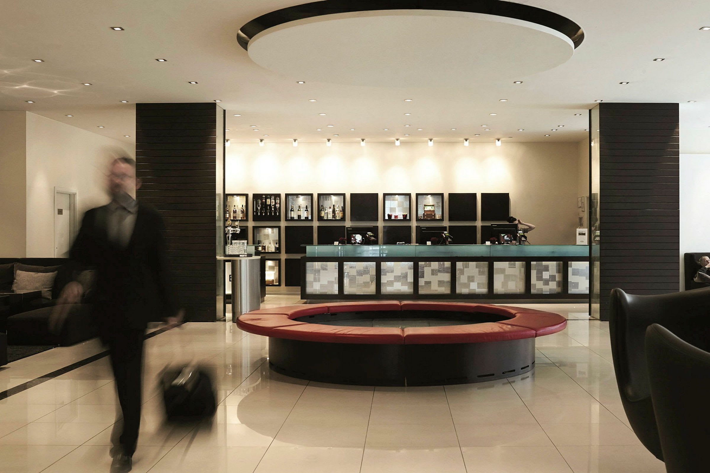 En godt opplyst hotellobby med en resepsjon, moderne sitteplasser og et rundt sofahjørne. En person med bagasje er i bevegelse. (AI-generert tekst)