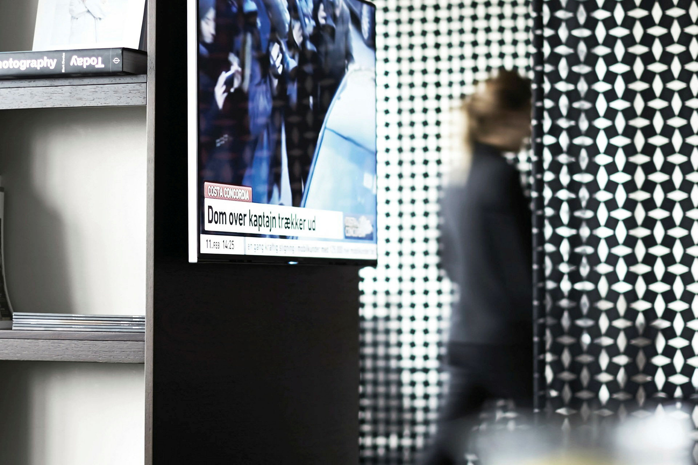 Et rom med en veggmontert TV som viser nyheter, en person går forbi i bakgrunnen med uskarphet. (AI-generert tekst)