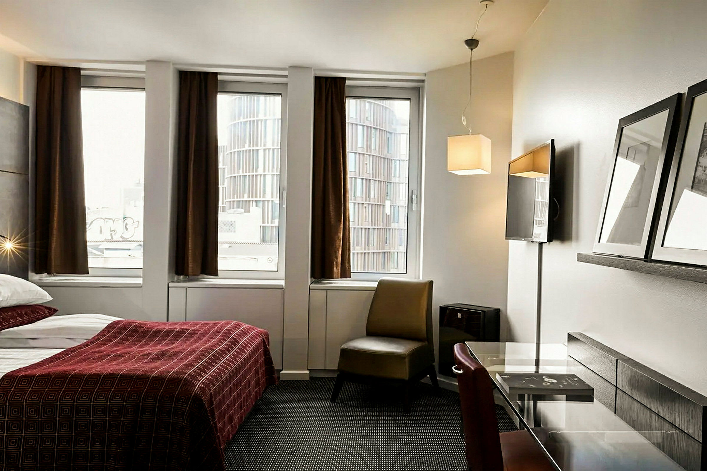 Et hotellrom med en seng med rødt sengeteppe, et skrivebord og en stol foran tre vinduer med brune gardiner. (AI-generert tekst)