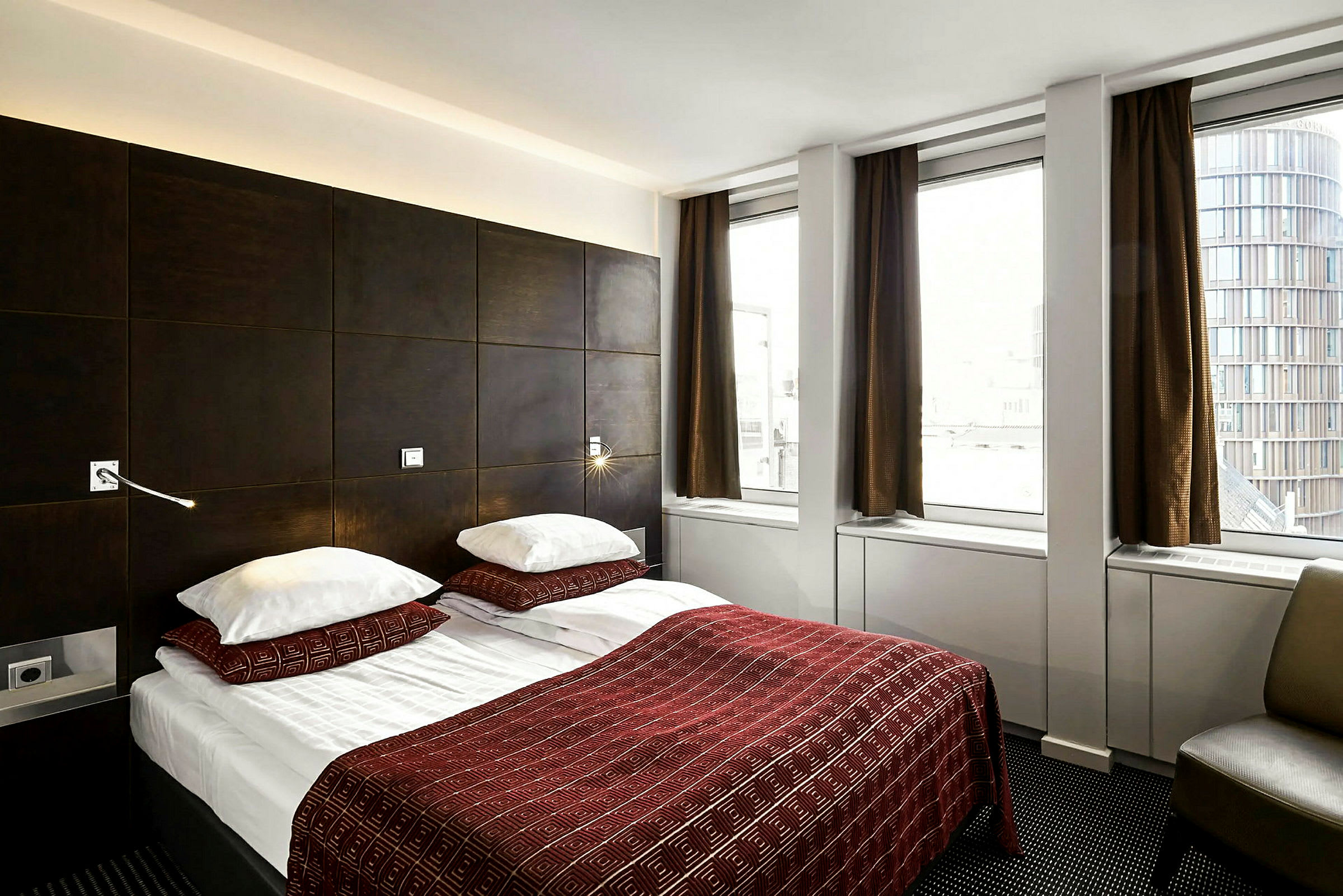 Et hotellrom med en dobbeltseng oppredd med rød dekorert sengeteppe og to puter. Rommet har mørke møbler og tre store vinduer med brune gardiner. (AI-generert tekst)