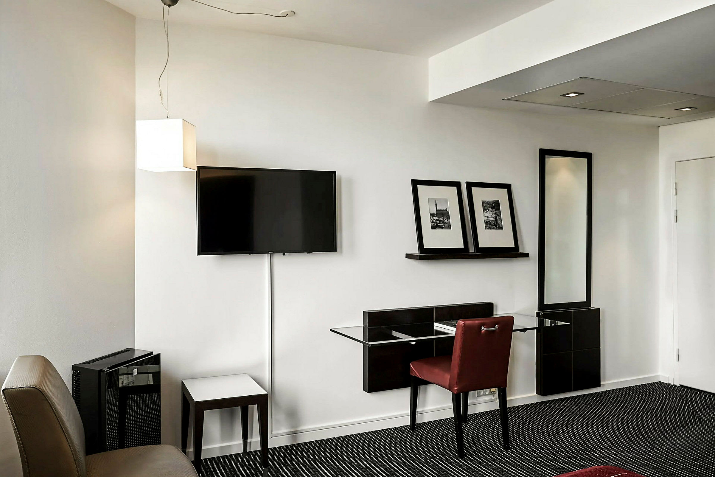 Et hotellrom med en vegg som har en veggmontert TV, en hylle med to innrammede bilder, og en sitteplass med et bord og en stol. (AI-generert tekst)