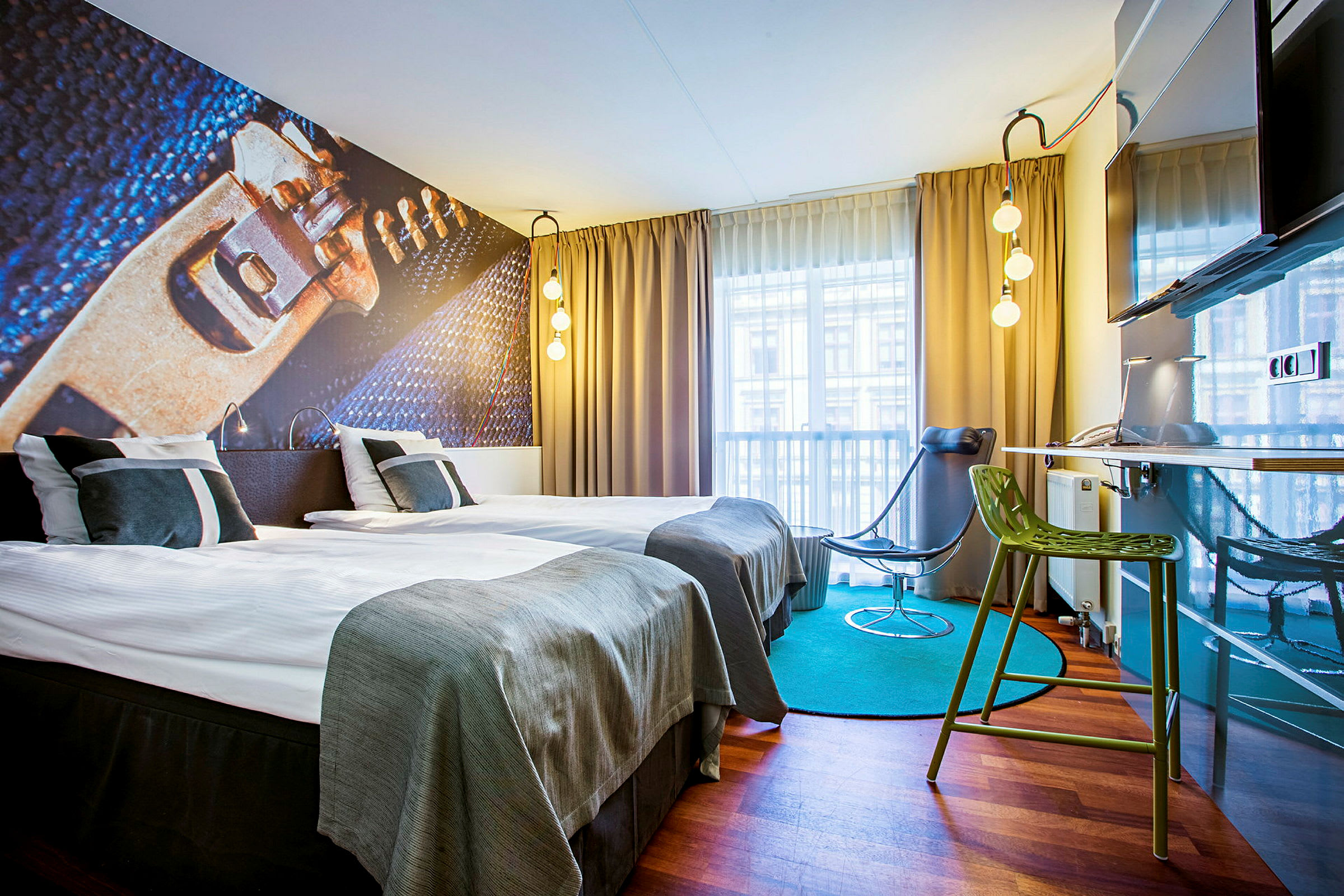 Et moderne designet hotellrom med to enkeltsenger, en illustrert veggdekorasjon, gardiner og vindusutsikt. Rommet har også en grønn stol, en svart stol og en veggmontert TV. (AI-generert tekst)
