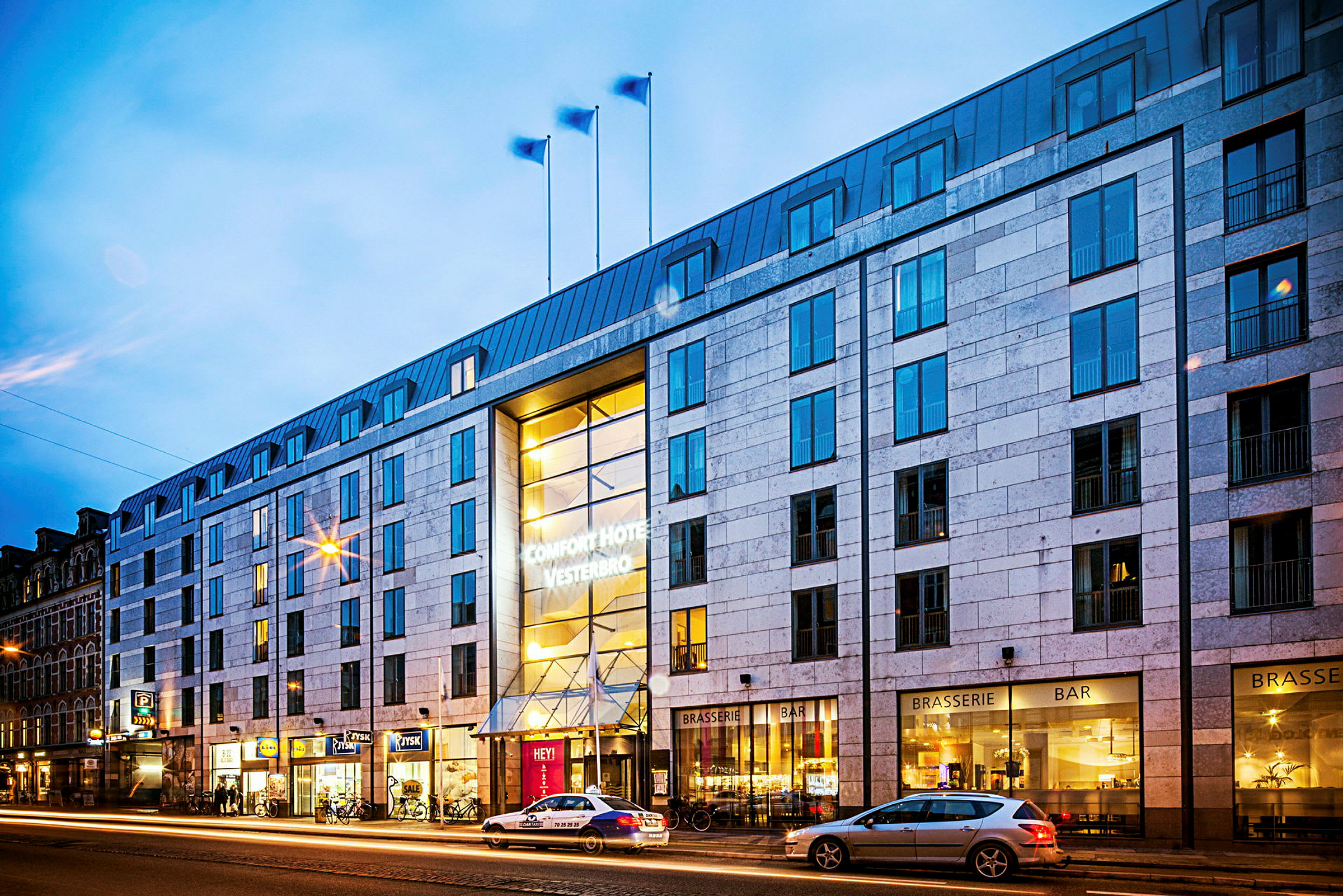 Eksteriør av hotellbygning om kvelden, med belysning og gaten med passerende biler. (AI-generert tekst)