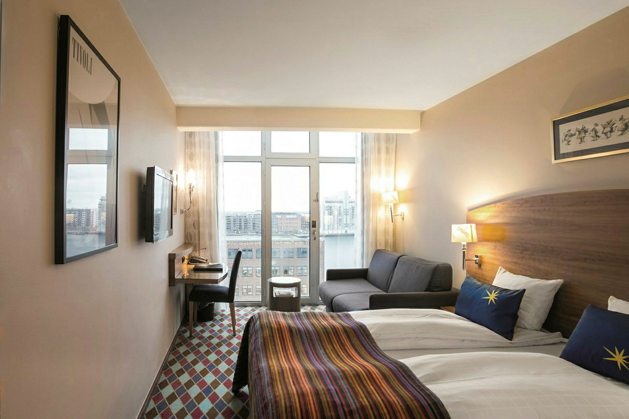 Et hotellrom med to enkeltsenger dekket med hvitt sengetøy og fargerike sengetepper. Det er også en grå sofa, et skrivebord med stol og utsikt over en by gjennom store vinduer. (AI-generert tekst)