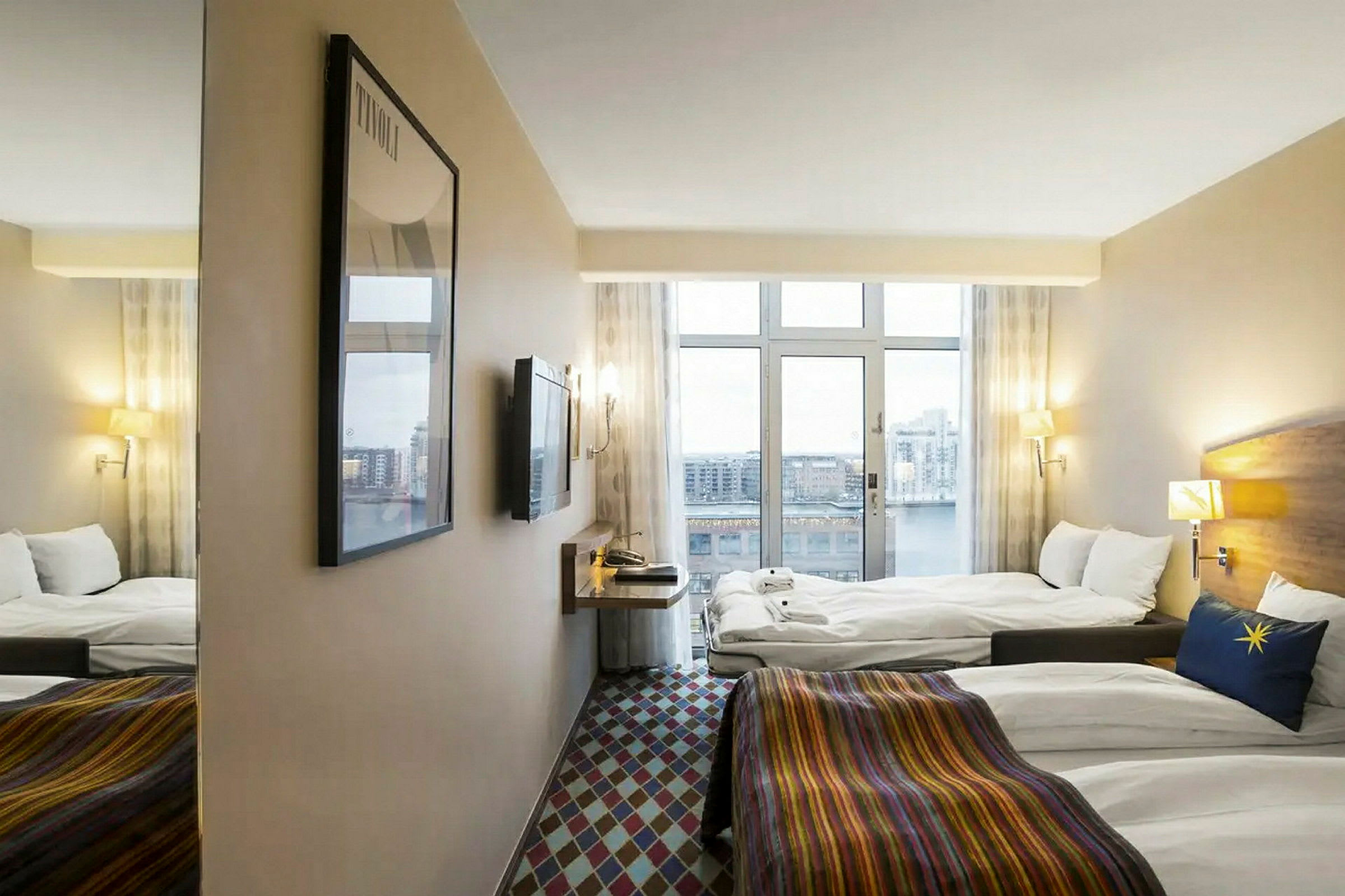Et hotellrom med vinduer som tilbyr utsikt over et bylandskap. Rommet er utstyrt med to senger, en veggmontert TV og et speil. (AI-generert tekst)