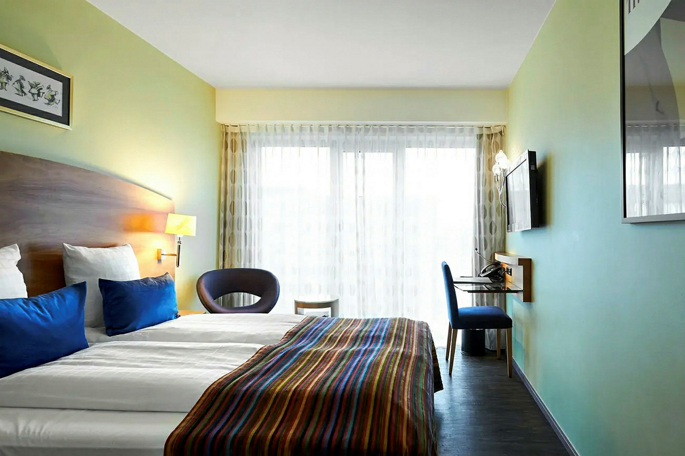 Et moderne hotellrom med en dobbeltseng, blå puter og et fargerikt sengeteppe. Rommet har også en veggmontert TV, et skrivebord og en stol, samt store vinduer med lyse gardiner. (AI-generert tekst)