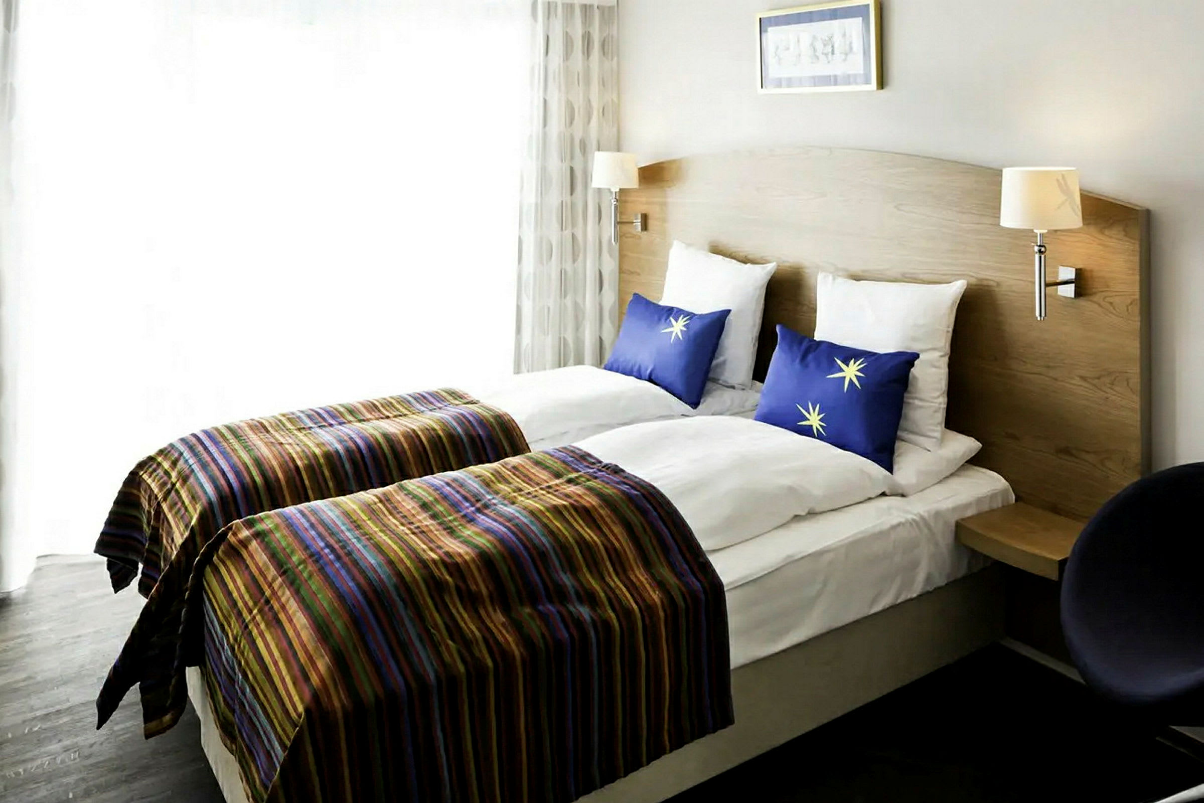 Hotellrom med to enkeltsenger med sengegavl i tre, stripete sengetepper og blå puter med stjernemotiv. (AI-generert tekst)