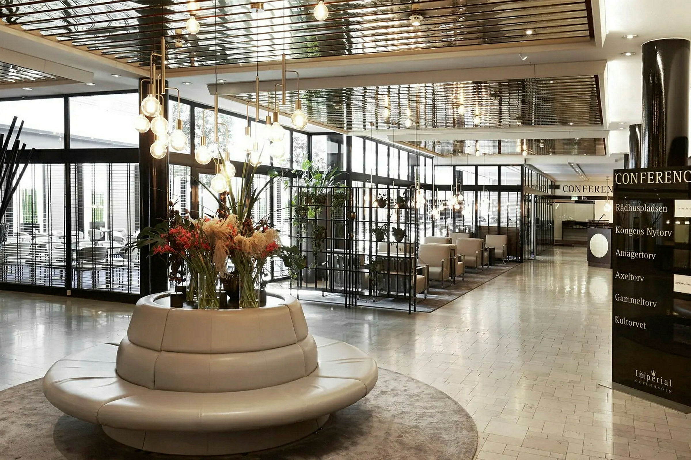 En moderne innredet hotellobby med en rund sittemøbel med blomsteroppsats i midten som står sentralt. I bakgrunnen sees flere sitteplasser og store glasspartier. (AI-generert tekst)