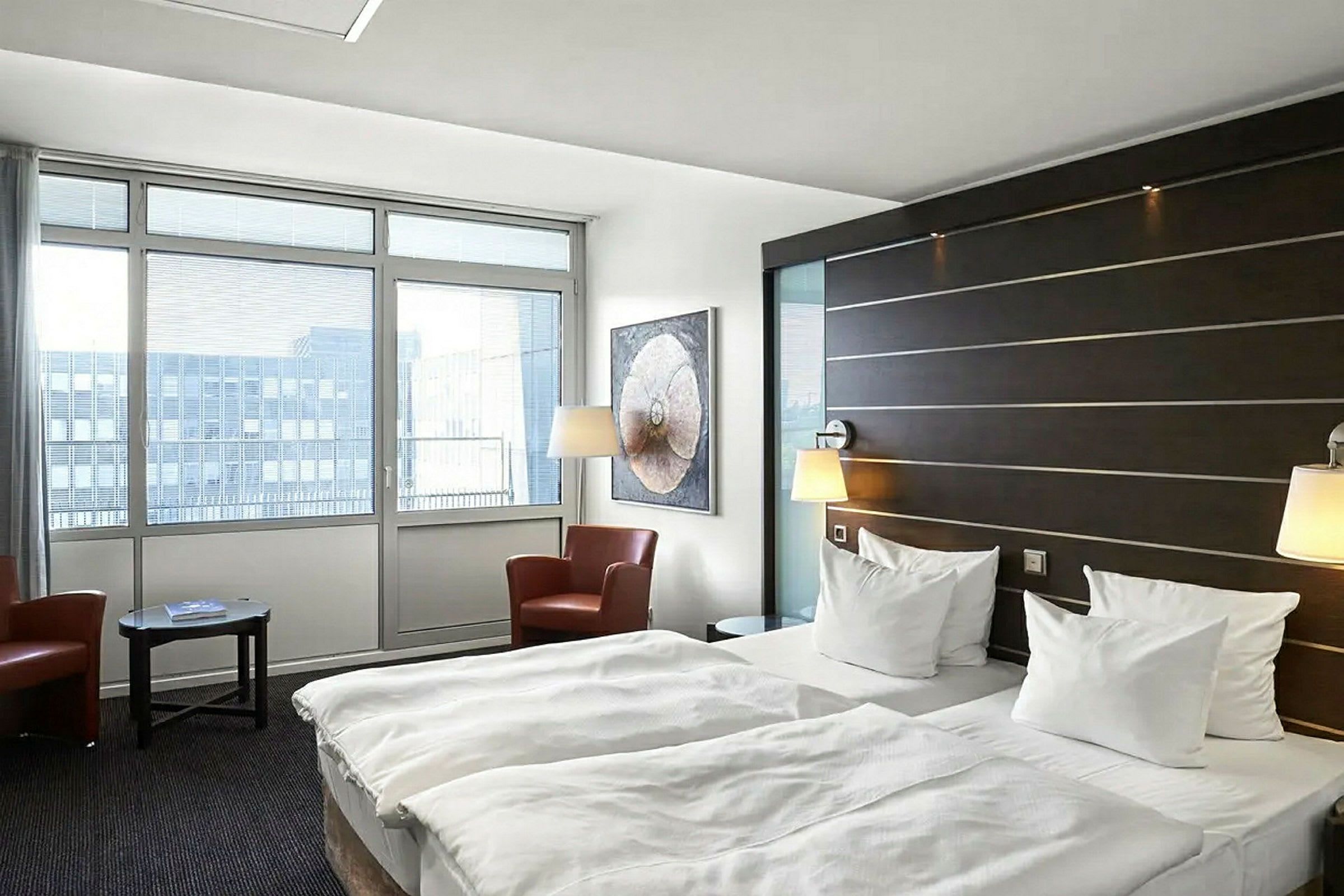 Et hotellrom med to enkeltsenger, en sittegruppe med to lenestoler og et bord samt store vinduer som slipper inn naturlig lys. (AI-generert tekst)