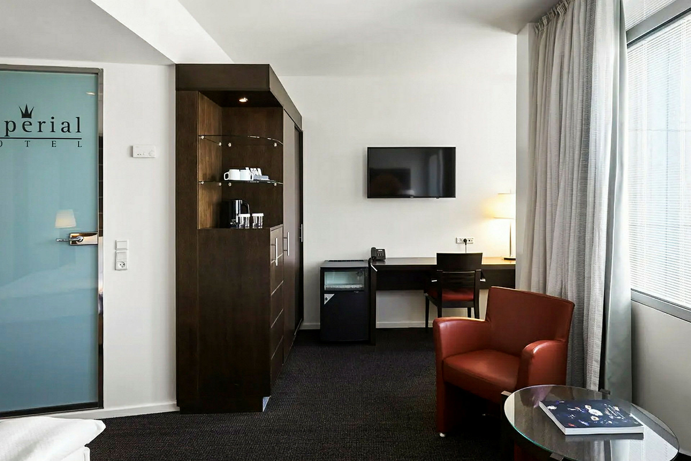 Et moderne hotellrom med møbler som et skrivebord, en rød lenestol og en TV. Det er også en minibar og en kaffestasjon. (AI-generert tekst)