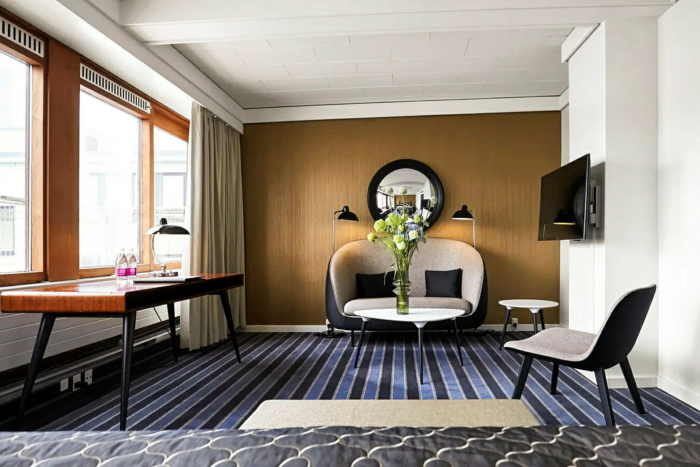 Et moderne hotellrom med en sofa, et bord og en TV, med store vinduer som slipper inn dagslys. (AI-generert tekst)