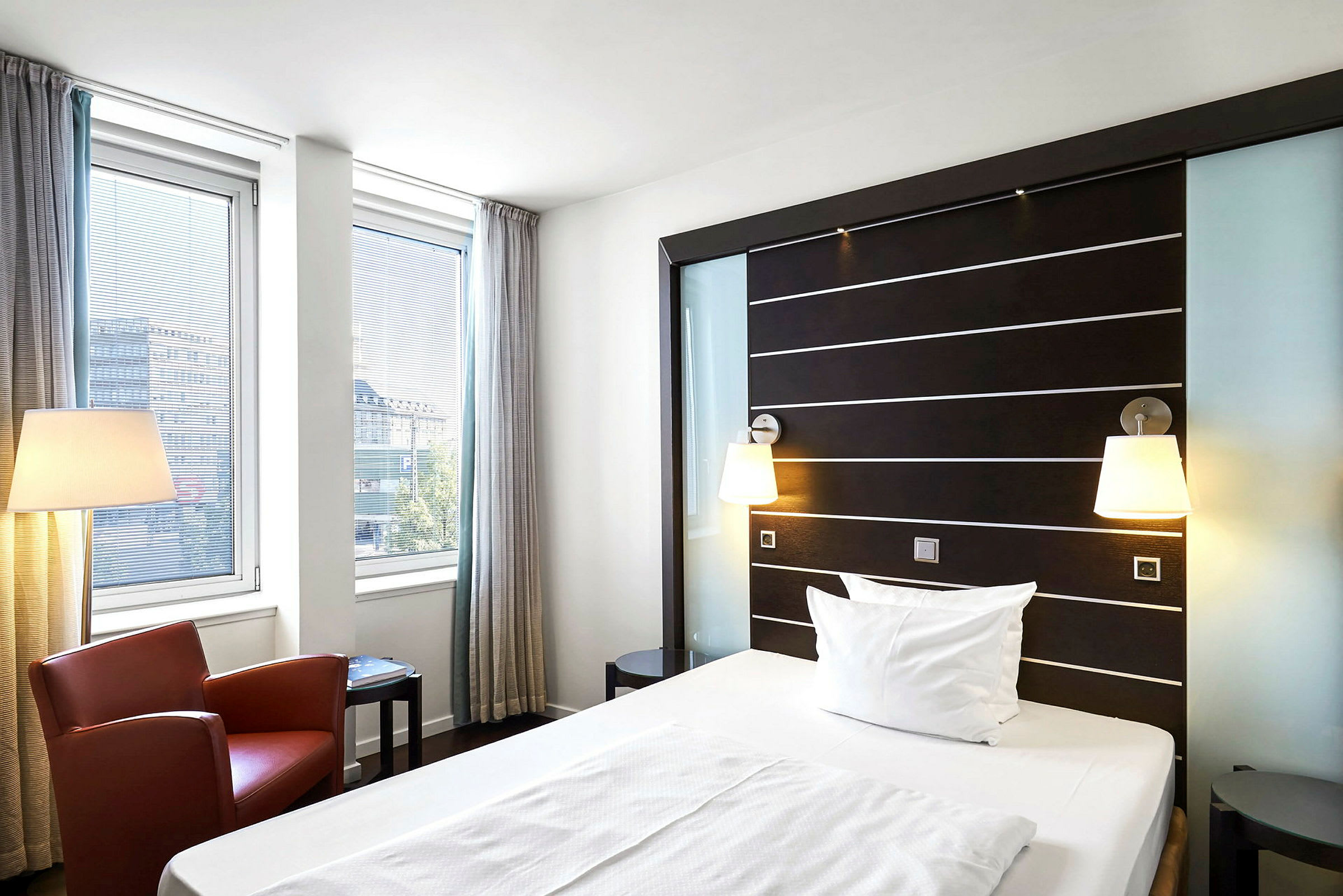 Et moderne hotellrom med en enkeltseng, to vinduer med gardiner, en rød lenestol og en gulvlampe. (AI-generert tekst)
