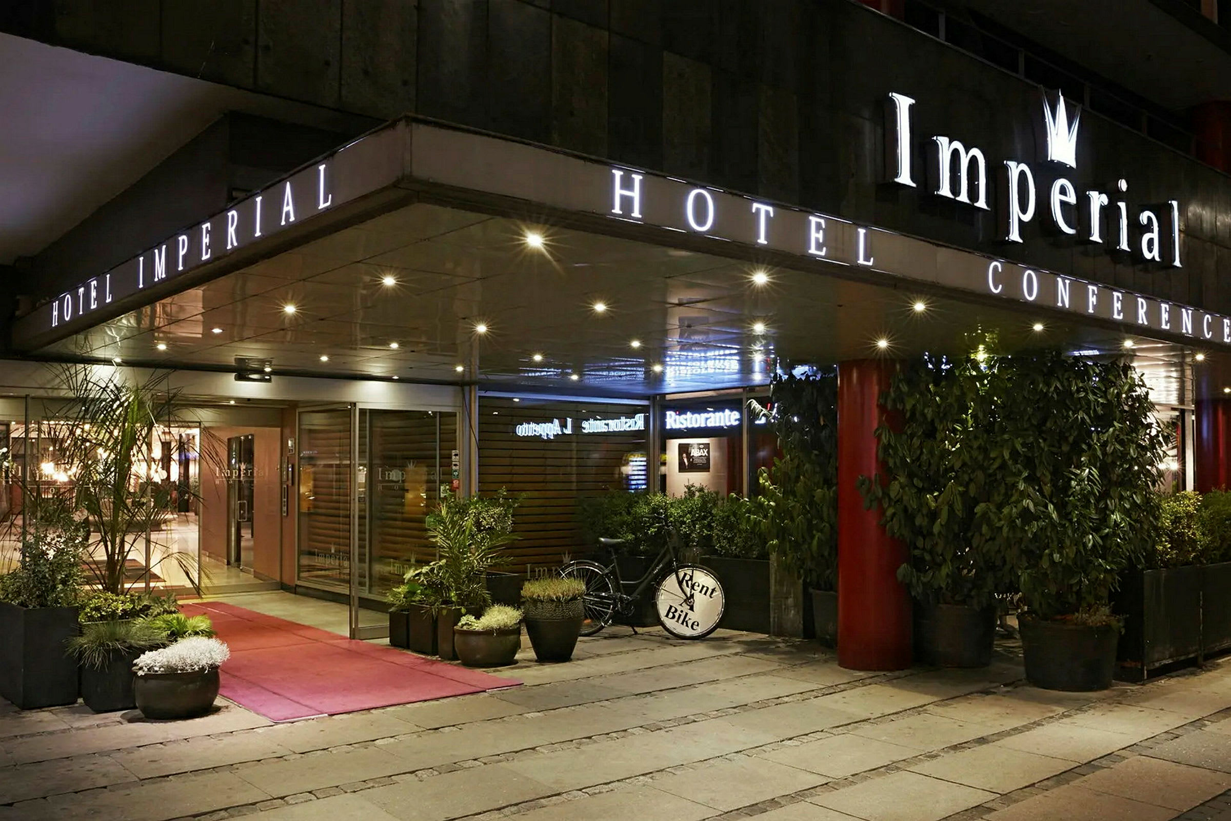 Inngangen til Hotel Imperial Conference med glassdører, planter i potter og en sykkel med skiltet 'Rent a Bike'. (AI-generert tekst)
