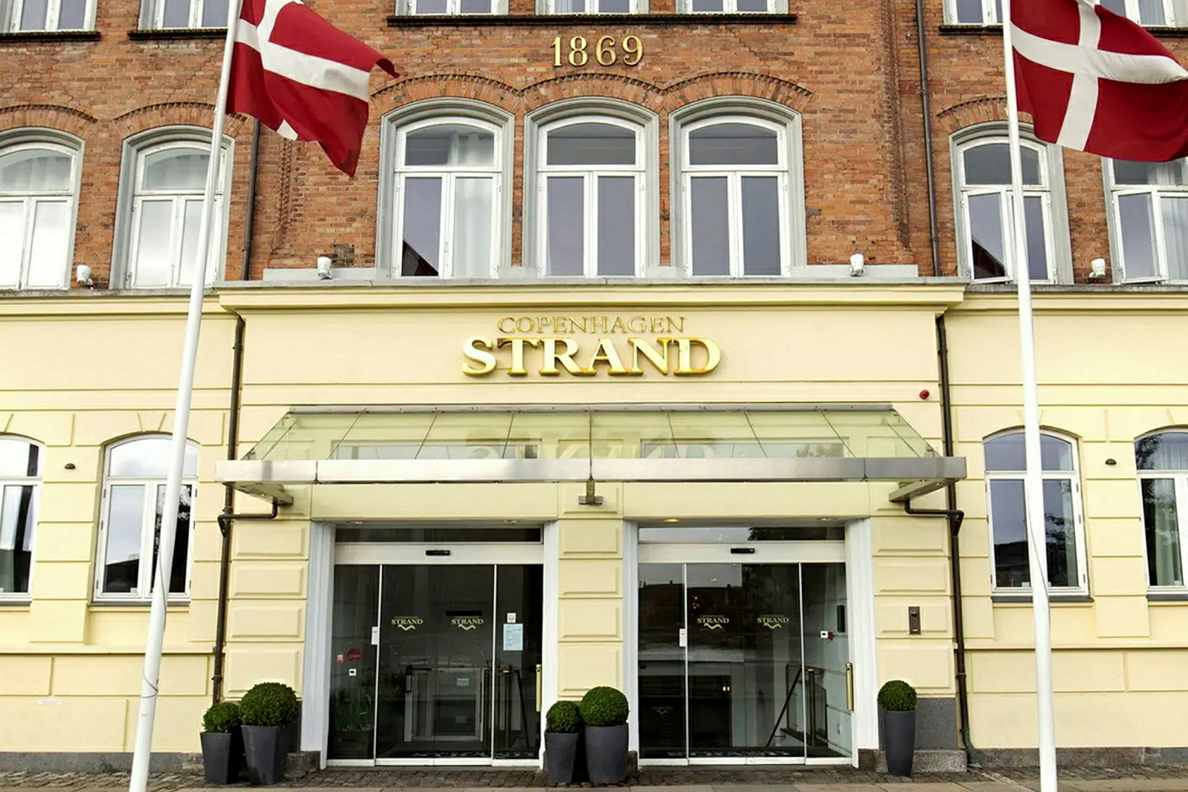 En bygning med gul fasade og et skilt der det står "Copenhagen Strand" over inngangsdørene. To danske flagg henger på hver side. (AI-generert tekst)