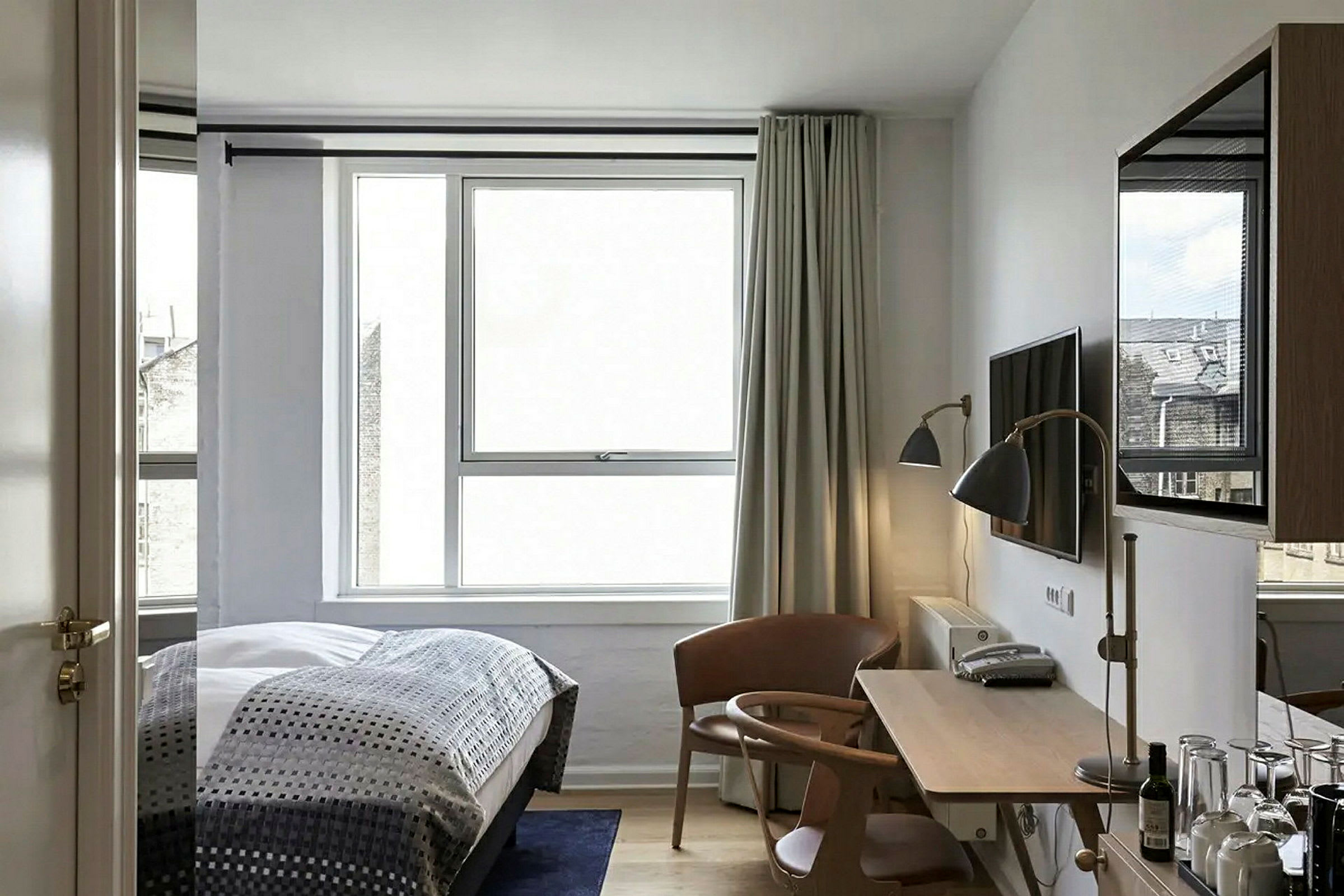 Bildet viser et moderne hotellrom med en seng, et skrivebord, en stol og et stort vindu. (AI-generert tekst)