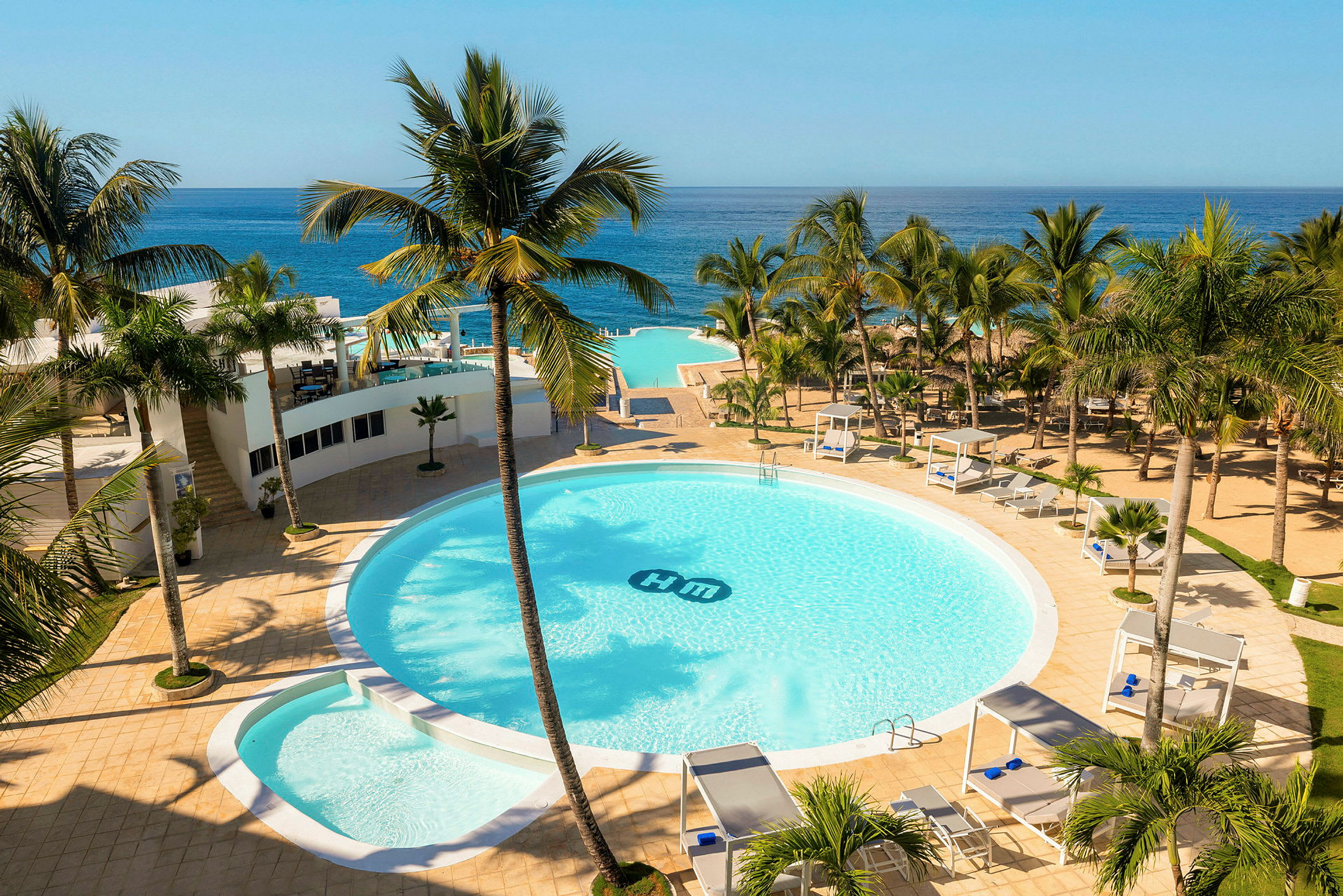 Et hotellområde med et stort rundt basseng omgitt av palmer, solsenger og cabanaer. Utsikt over havet i bakgrunnen. (AI-generert tekst)
