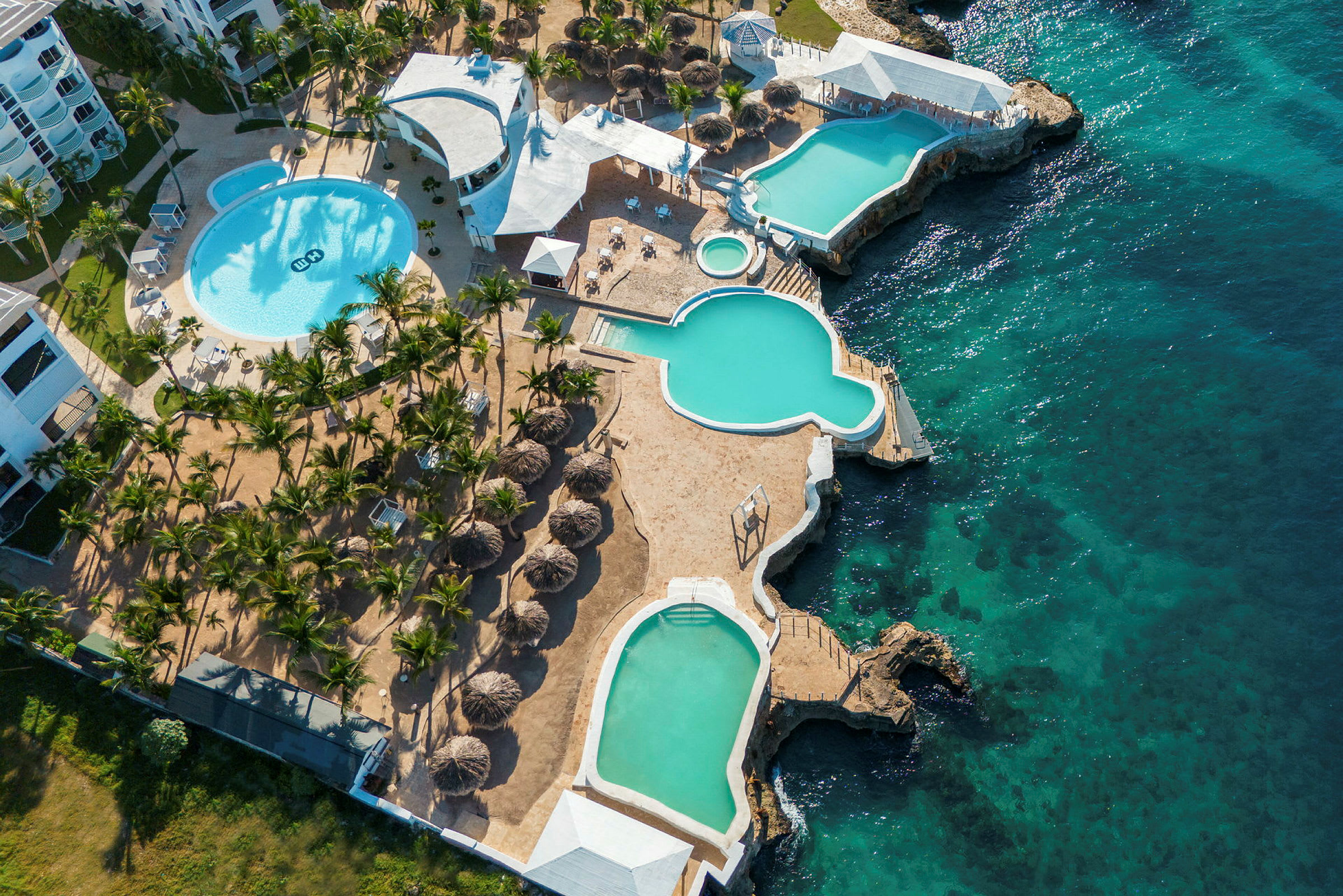 Flyfoto av et resort med flere bassenger, omgitt av solsenger og palmer, ved siden av et klart blått hav. (AI-generert tekst)