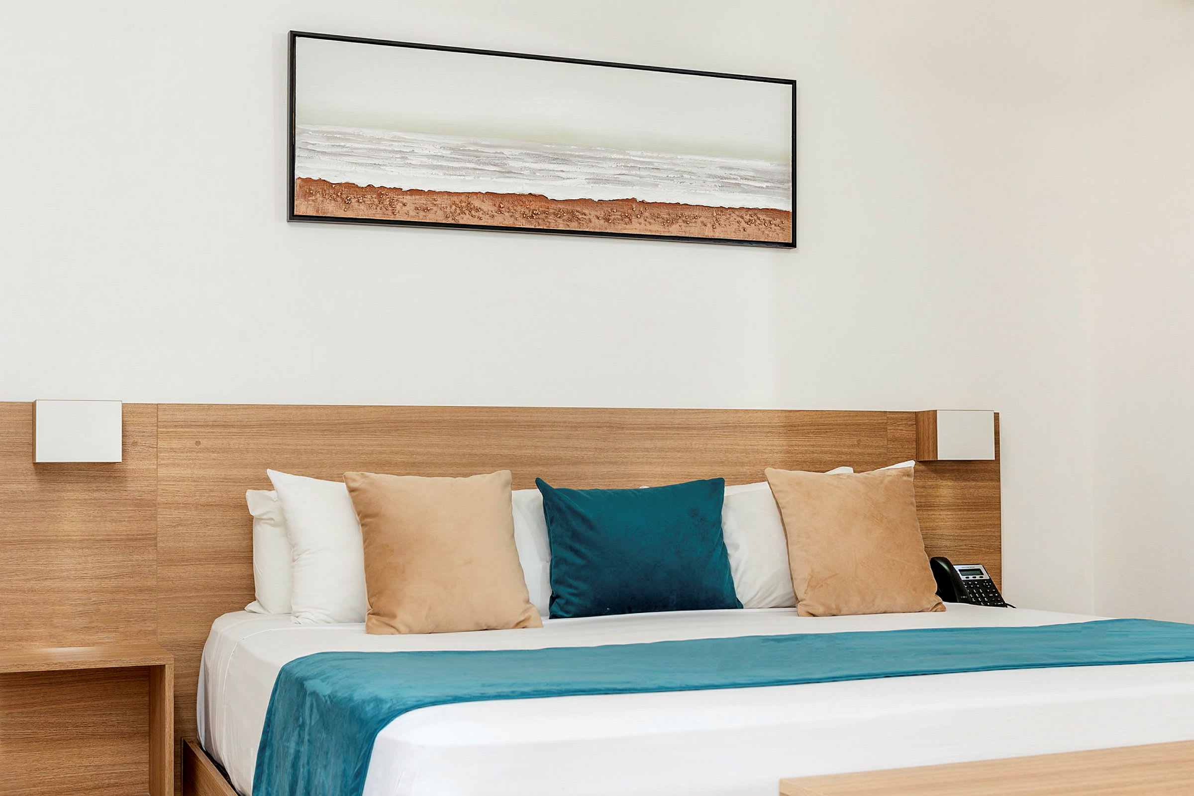 Et hotellrom med en seng med puter i beige og blå farger samt et blått sengeteppe. Et maleri med et landskapsmotiv henger på veggen over. (AI-generert tekst)