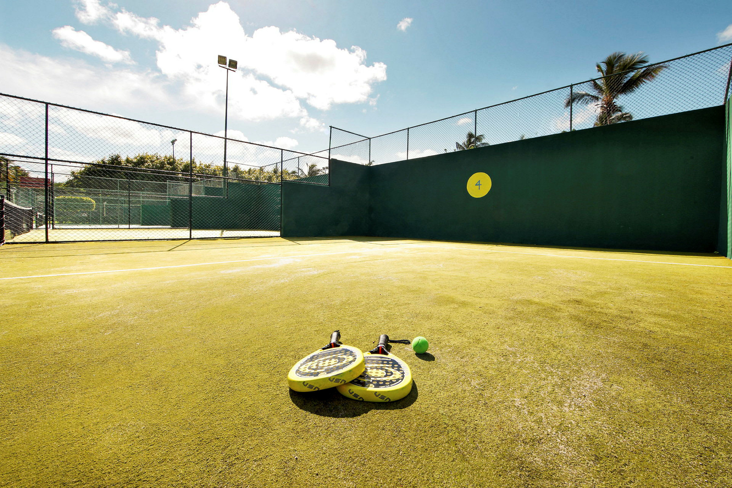 En padelbane med gule racketer og en ball plassert på bakken. Solfylt vær. (AI-generert tekst)