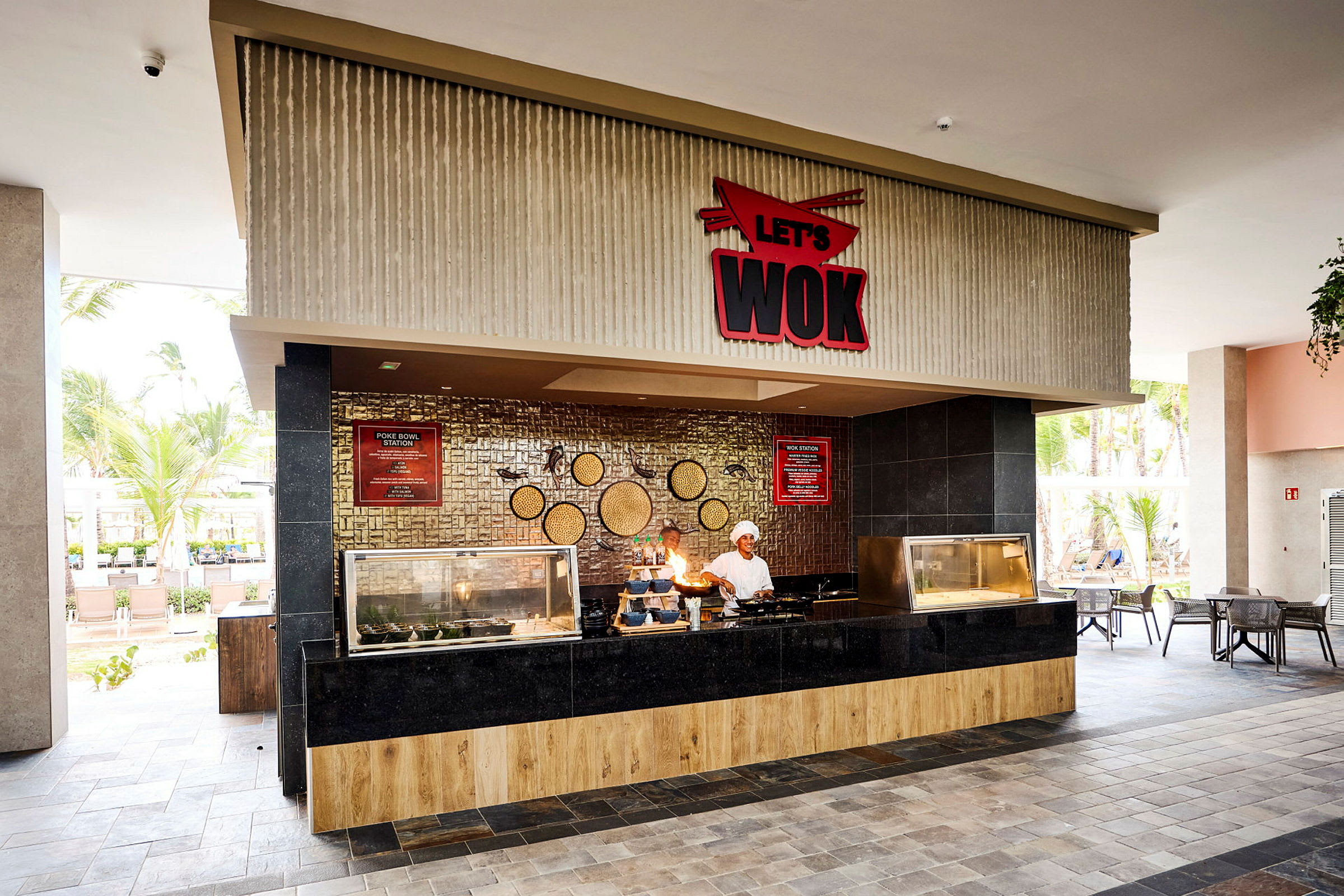 Bilde av en restaurant med en serveringsdisk merket 'Let's Wok'. Interiøret har en moderne design med tre og svarte paneler, og det er utendørs omgivelser med palmer i bakgrunnen. (AI-generert tekst)