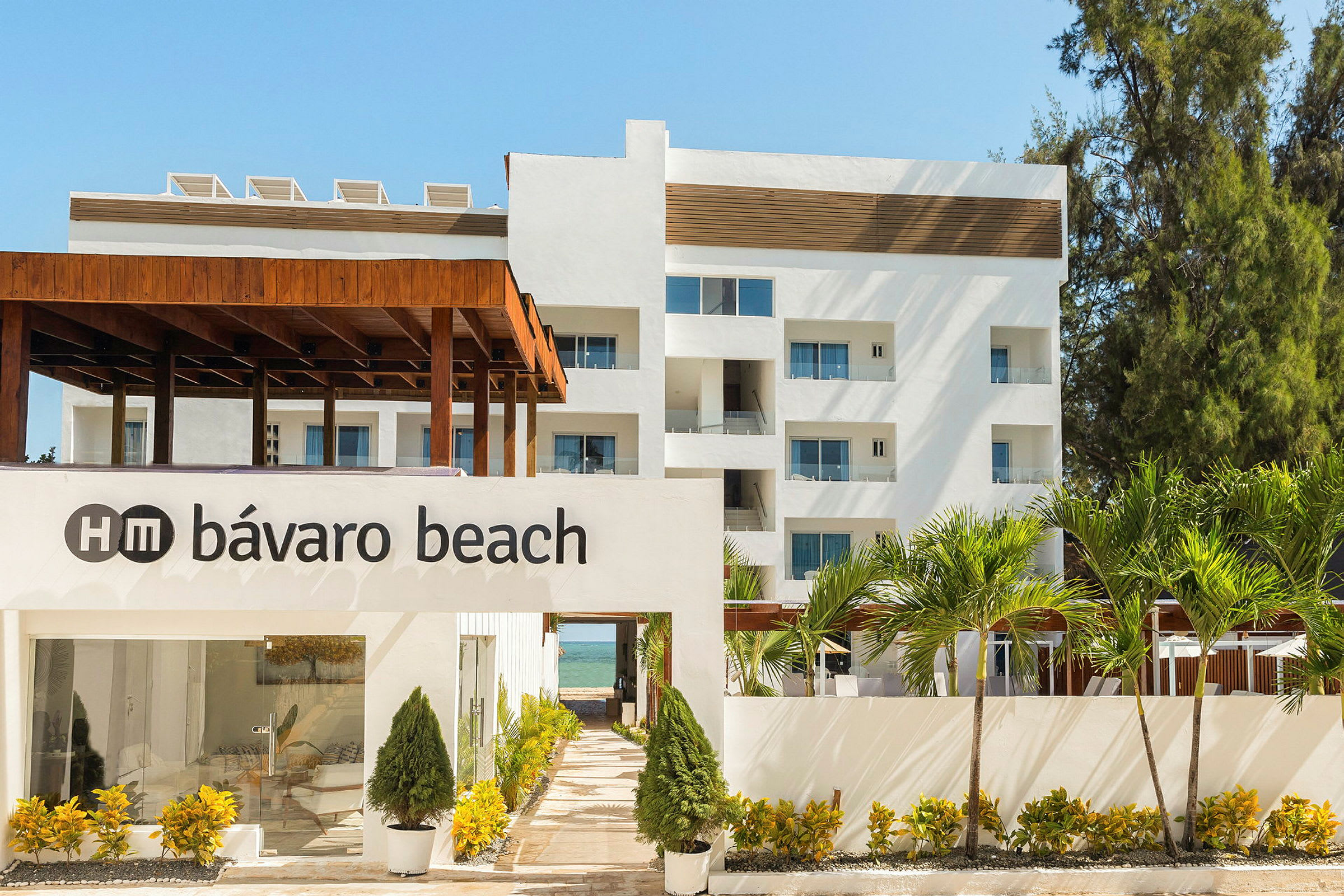 Bilde av en hvit bygning med teksten "HM Bávaro Beach" på en vegg foran, omgitt av frodig vegetasjon og palmer. (AI-generert tekst)