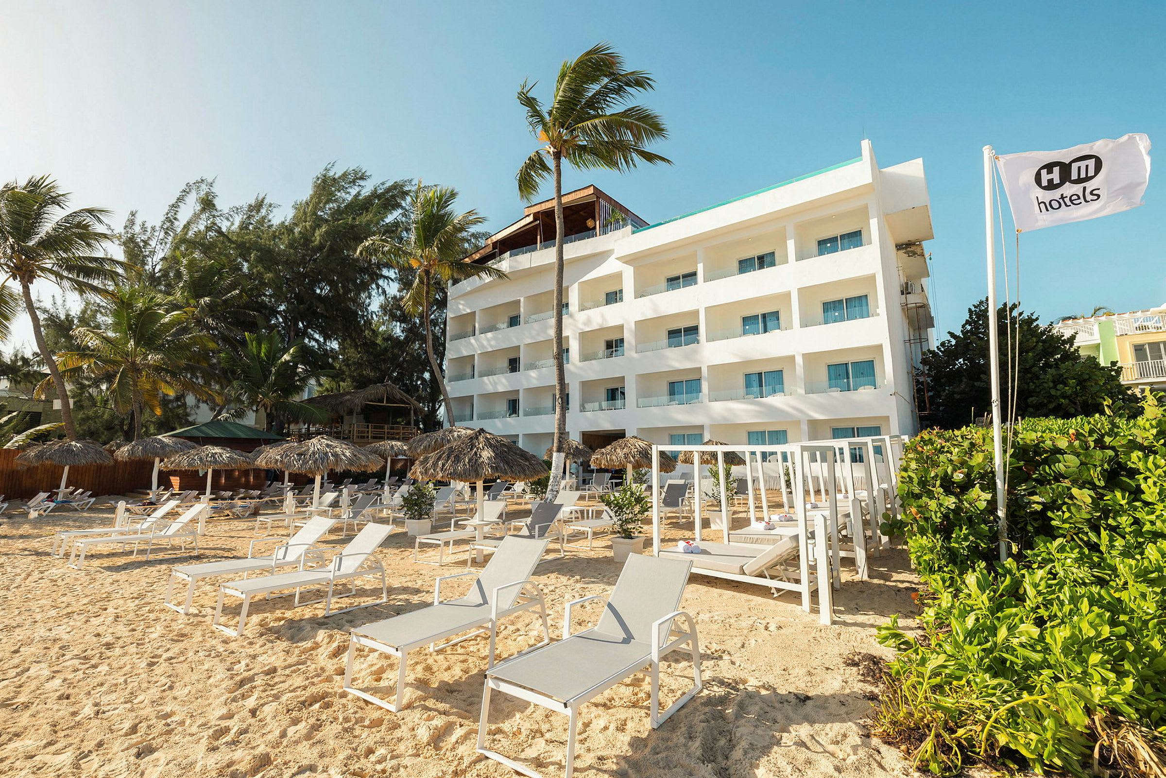 En hvit hotellbygning ved en sandstrand omgitt av parasoller og solsenger. (AI-generert tekst)