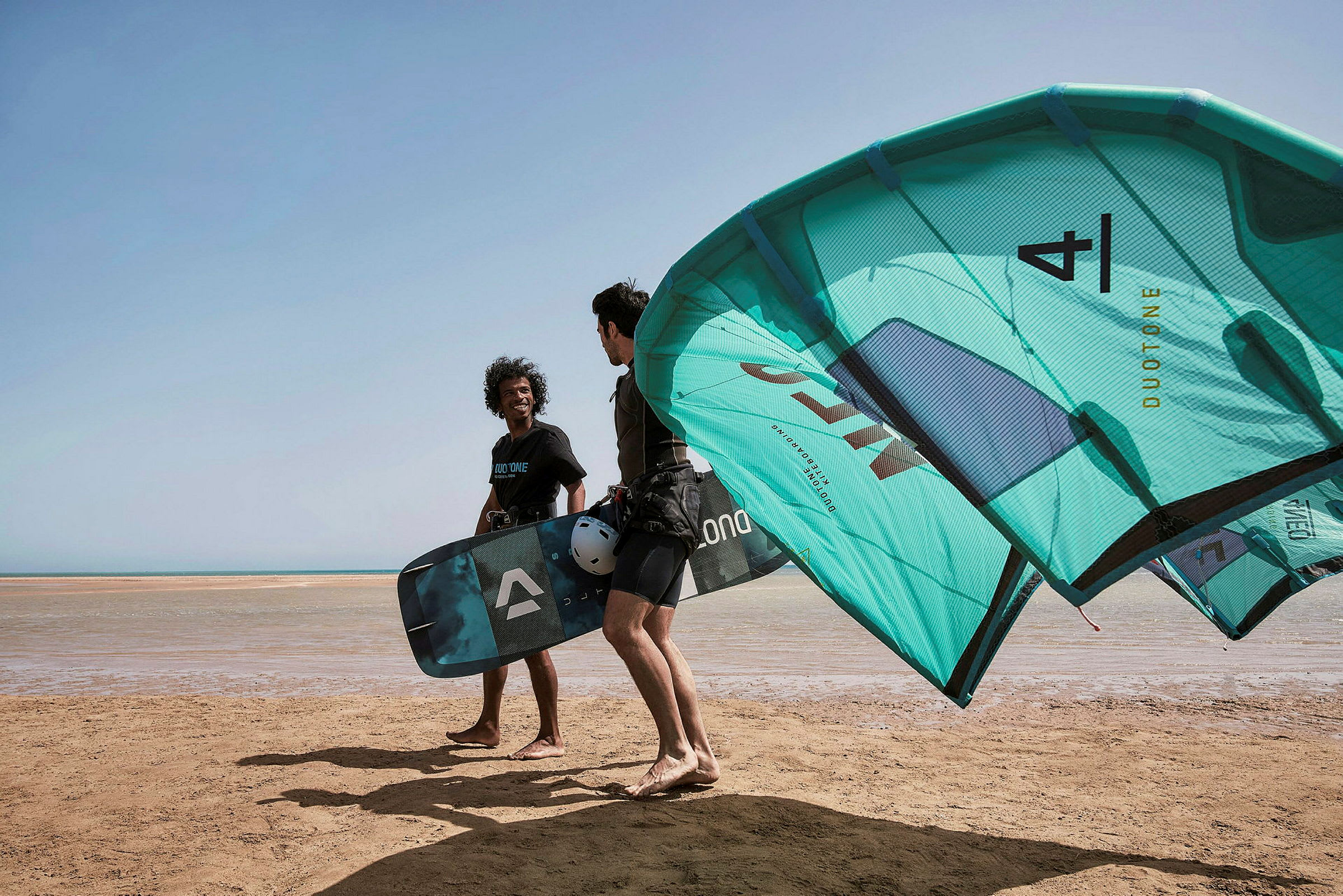 To personer med kiteutstyr står på en strand. (AI-generert tekst)