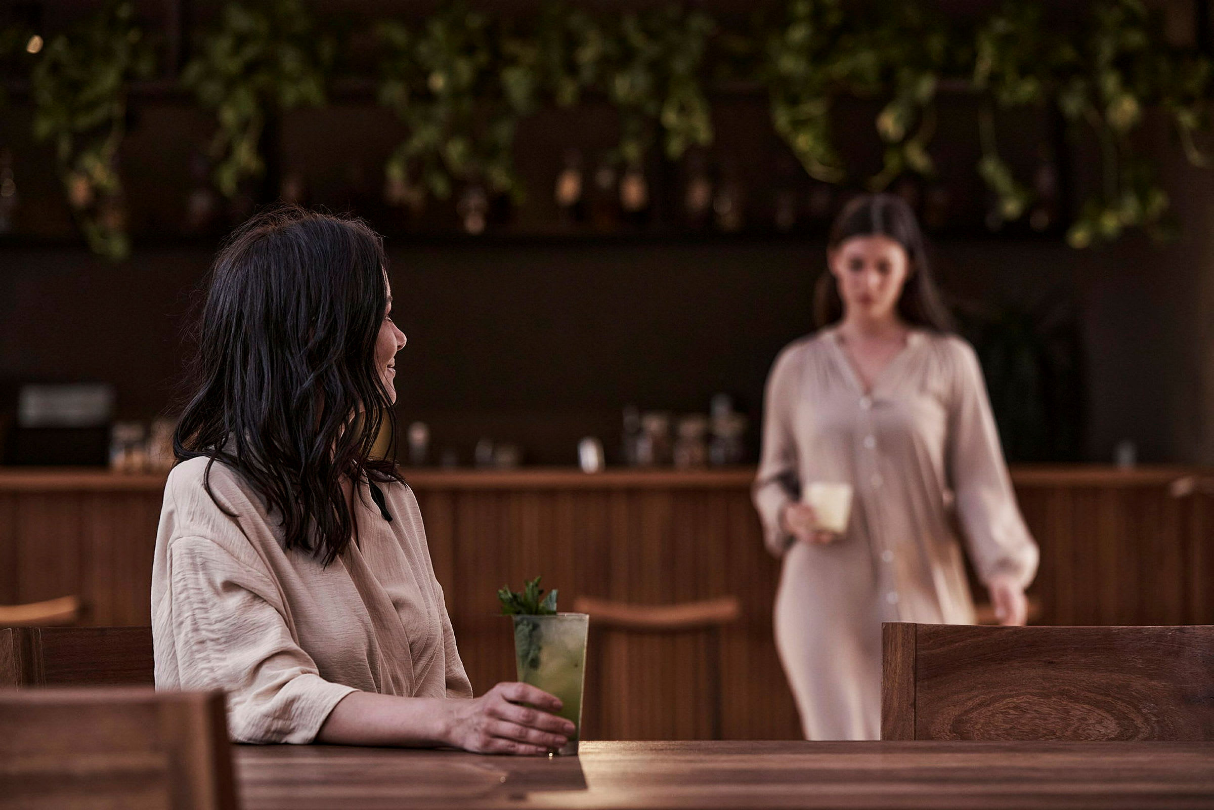En person holder en drink ved et bord med en bar i bakgrunnen. (AI-generert tekst)