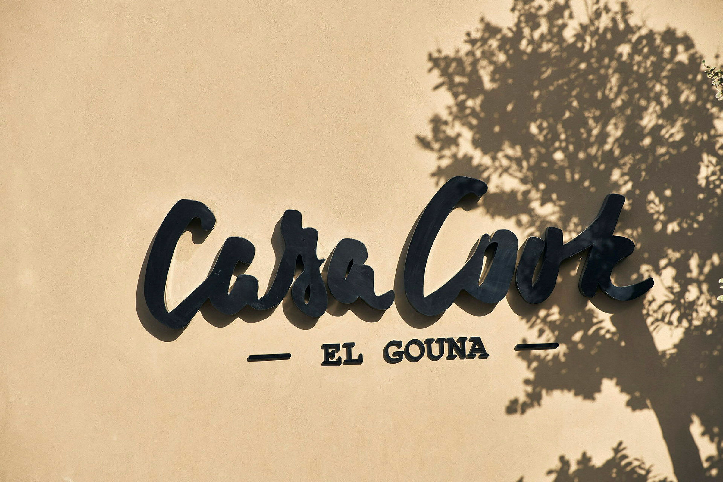 Logoen for Casa Cook El Gouna på en vegg, med skyggen av et tre. (AI-generert tekst)