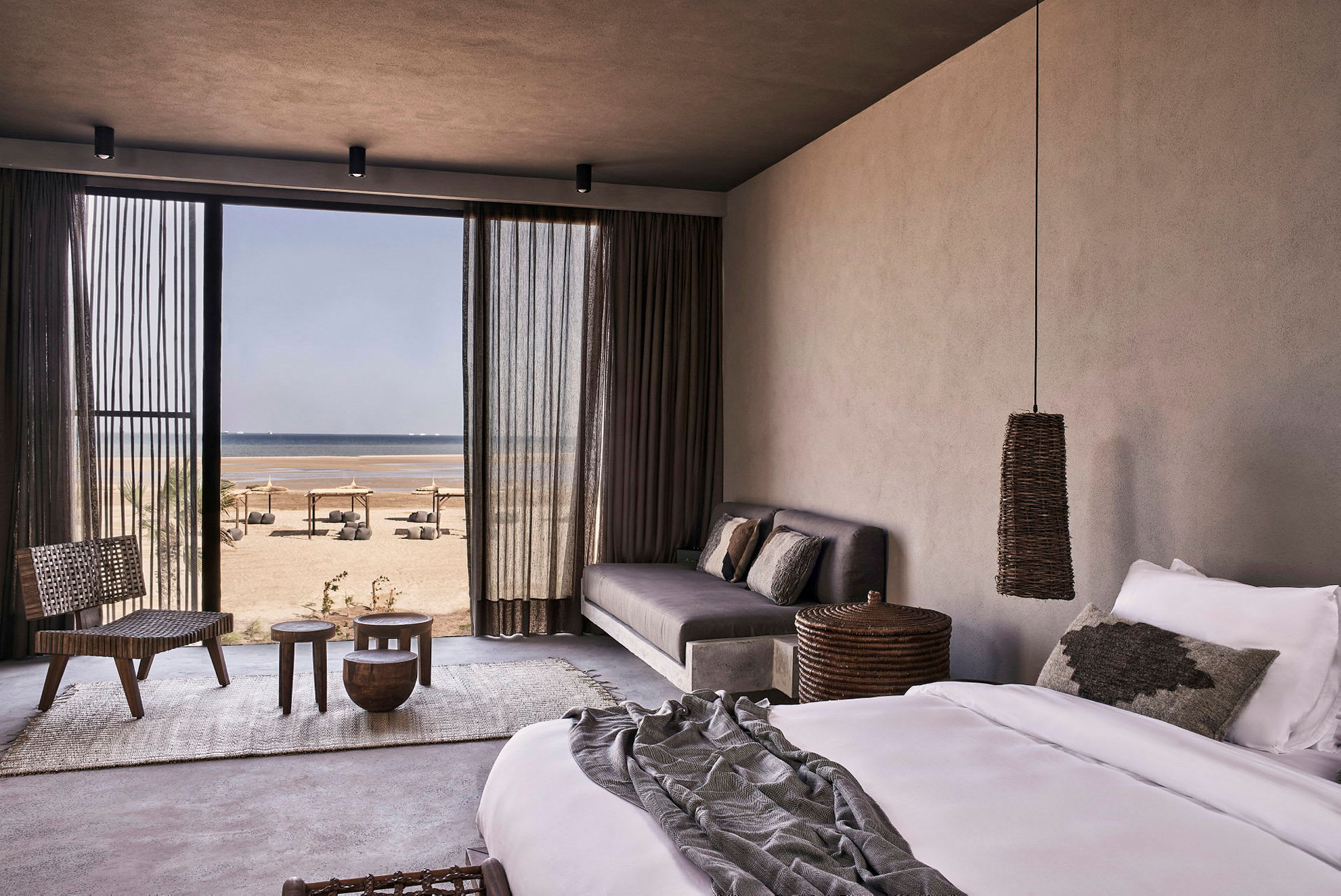 Et hotellrom med seng, sofa og utsikt mot stranden gjennom en stor glassdør. (AI-generert tekst)