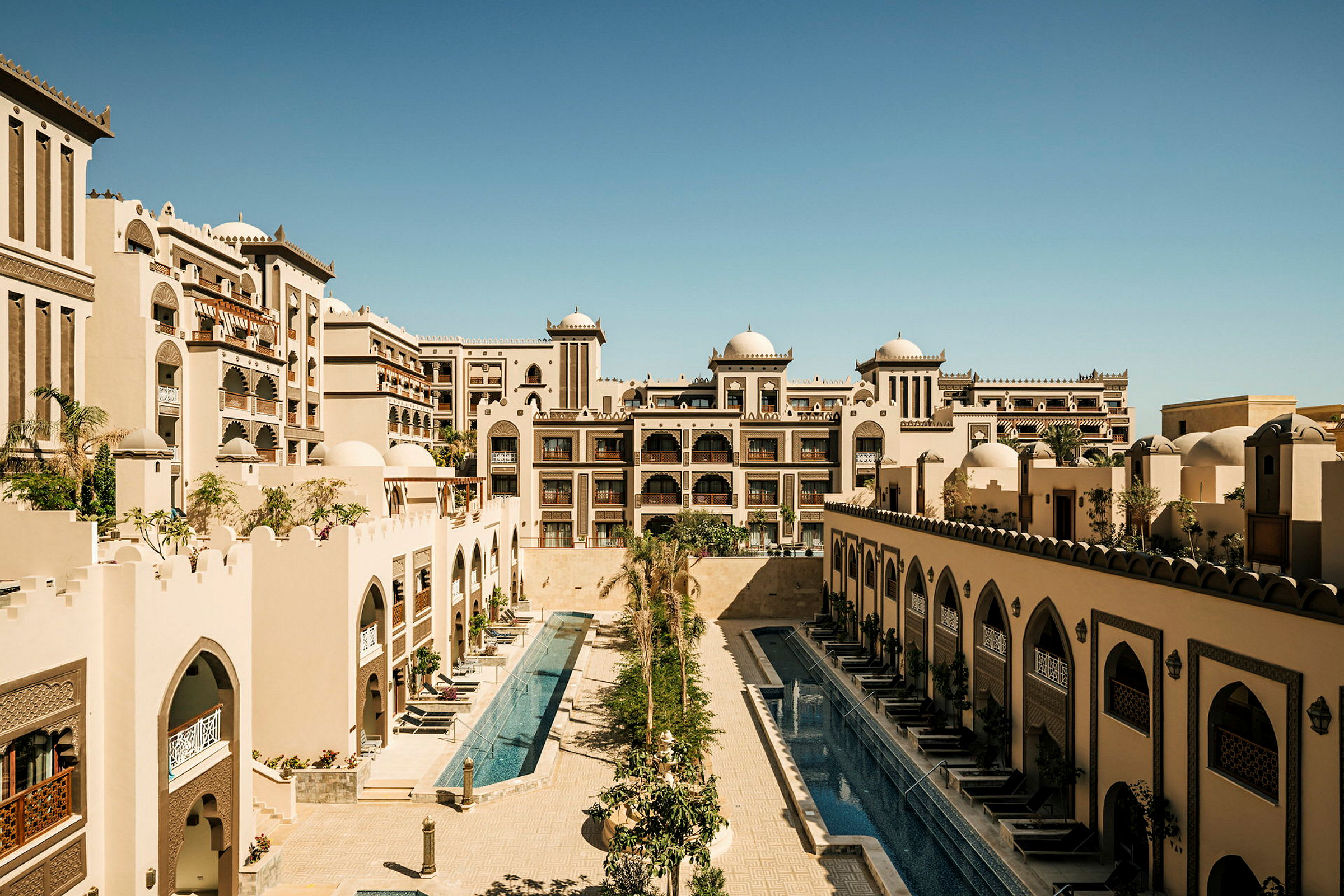Bildet viser et hotellkompleks med arkitektur inspirert av arabisk stil, inkludert kupler og ornamenterte buer, samt to langstrakte bassenger omgitt av bygninger. (AI-generert tekst)
